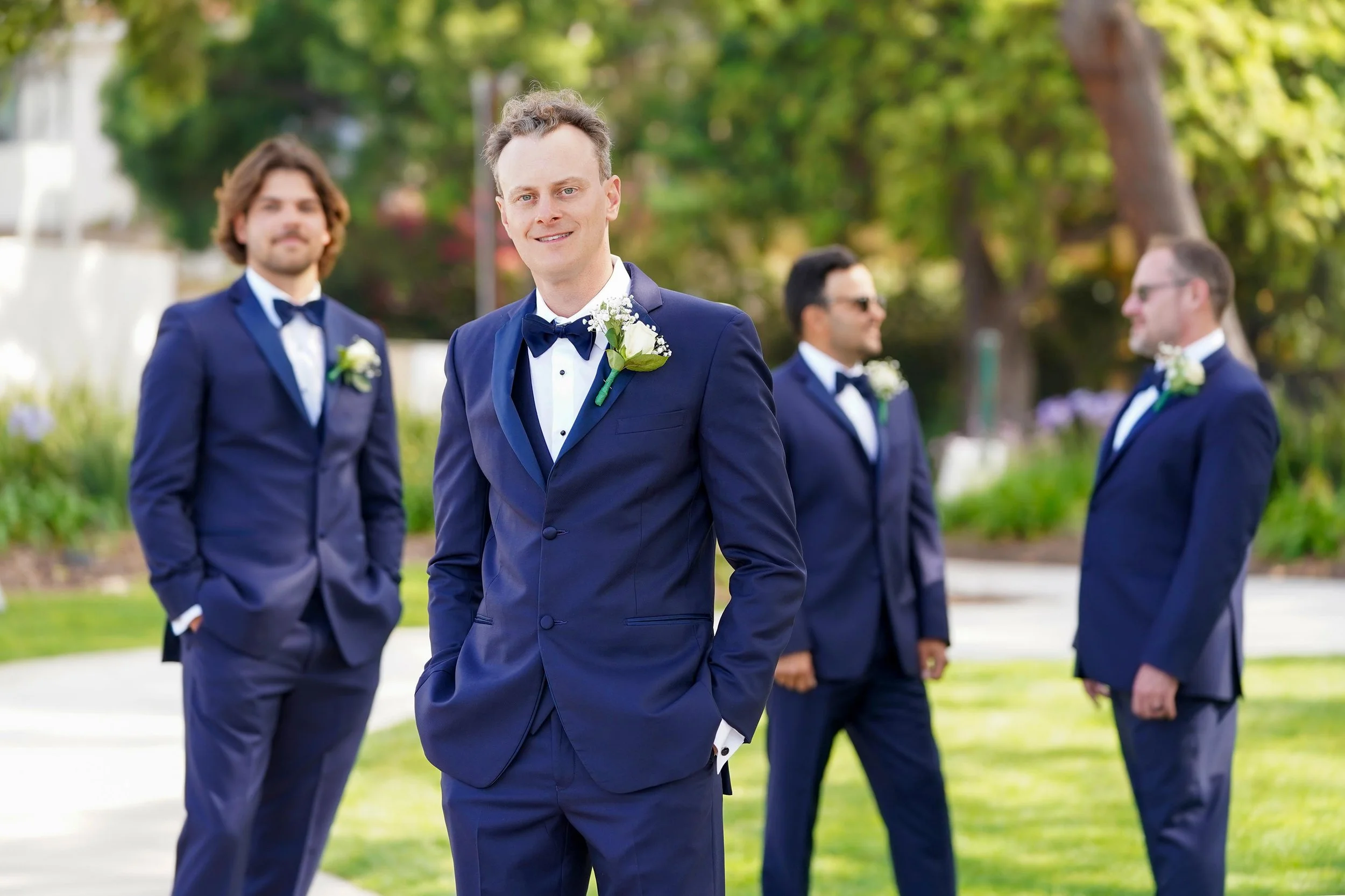 A groom in a navy blue tuxedo with a black shawl lapel and black bow tie stands smiling at the camera, flanked by three groomsmen wearing matching navy blue suits and bow ties in an outdoor park or garden setting.