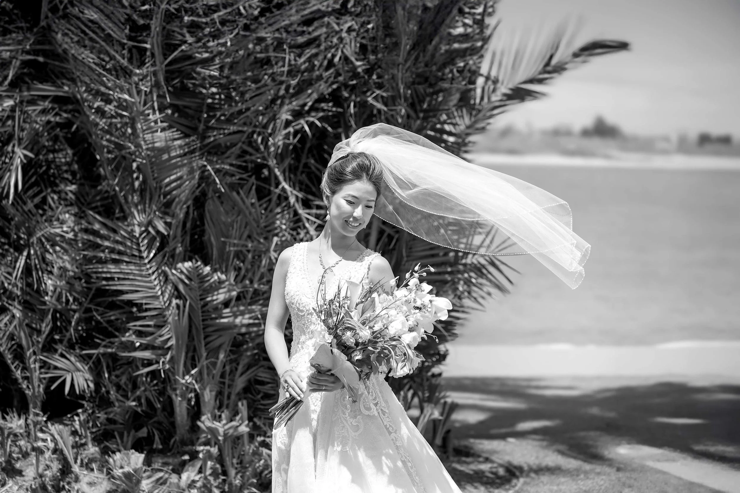 A black and white photograph of a smiling bride in a white lace wedding gown and a flowing veil, holding a large bouquet of flowers outdoors with palm tree foliage and a view of the water in the background.