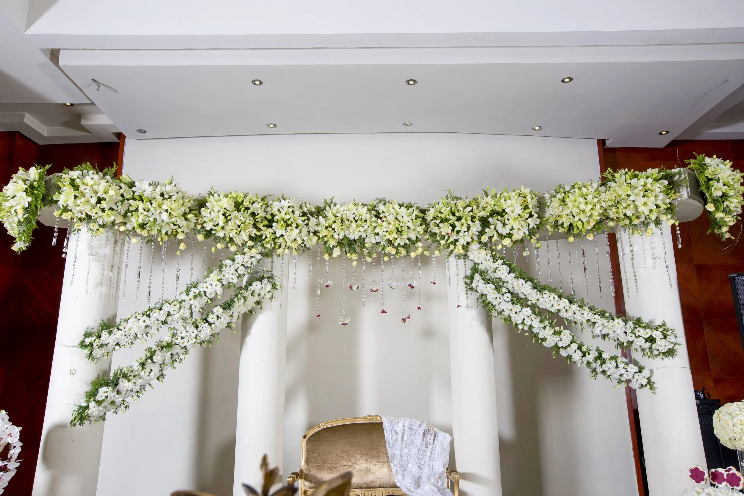 An elaborate indoor event display featuring a large horizontal swag of white flowers and hanging crystal accents, with additional white flower arrangements draped down to form an arch around a gold chair in a wall niche.