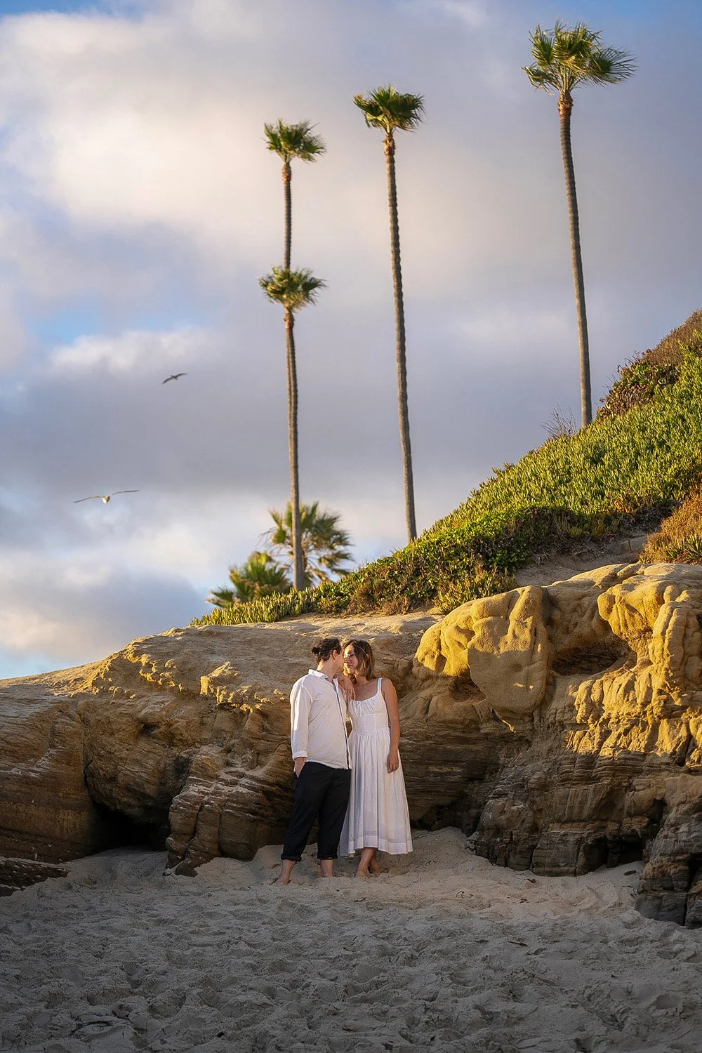 An outdoor photograph of a couple embracing and about to kiss on a sandy beach below a rocky bluff. The man wears a white long-sleeved shirt and dark pants, and the woman wears a white sundress; both are barefoot. Several tall palm trees grow from th