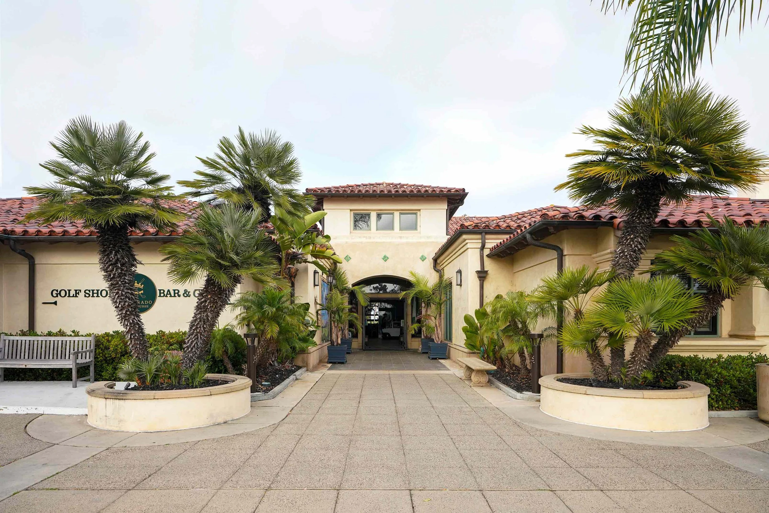 A single-story, mission-style building with a red tile roof features an arched entryway, text for "GOLF SHOP" and "BAR & GRILL," and is surrounded by numerous tall and short palm trees in Coronado, potted fan palms on a paved stone walkway.


