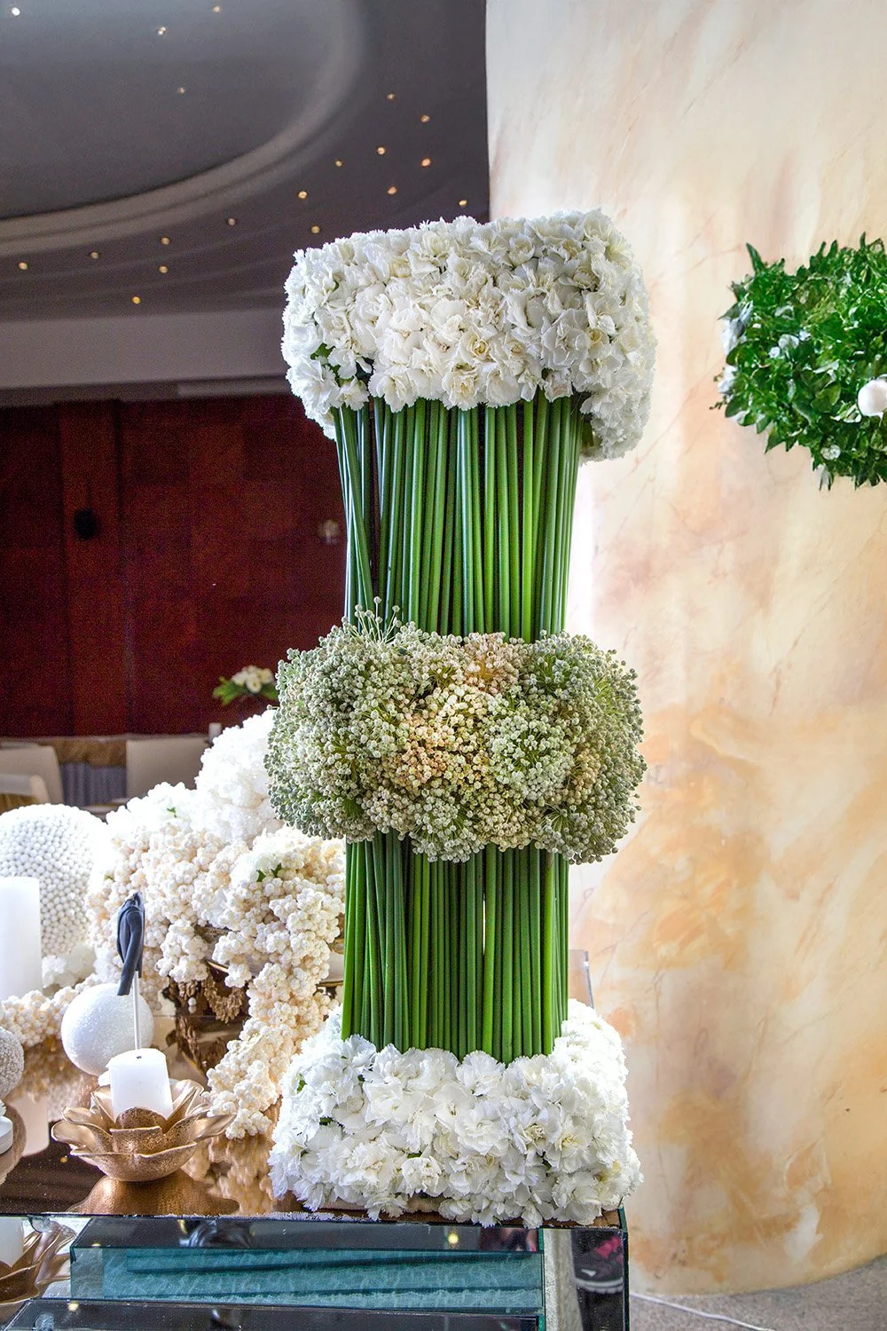 A tall, modern floral arrangement featuring vertical green stems flanked by bands of white and light green flowers, sitting on a mirrored table with additional white decor and candles.