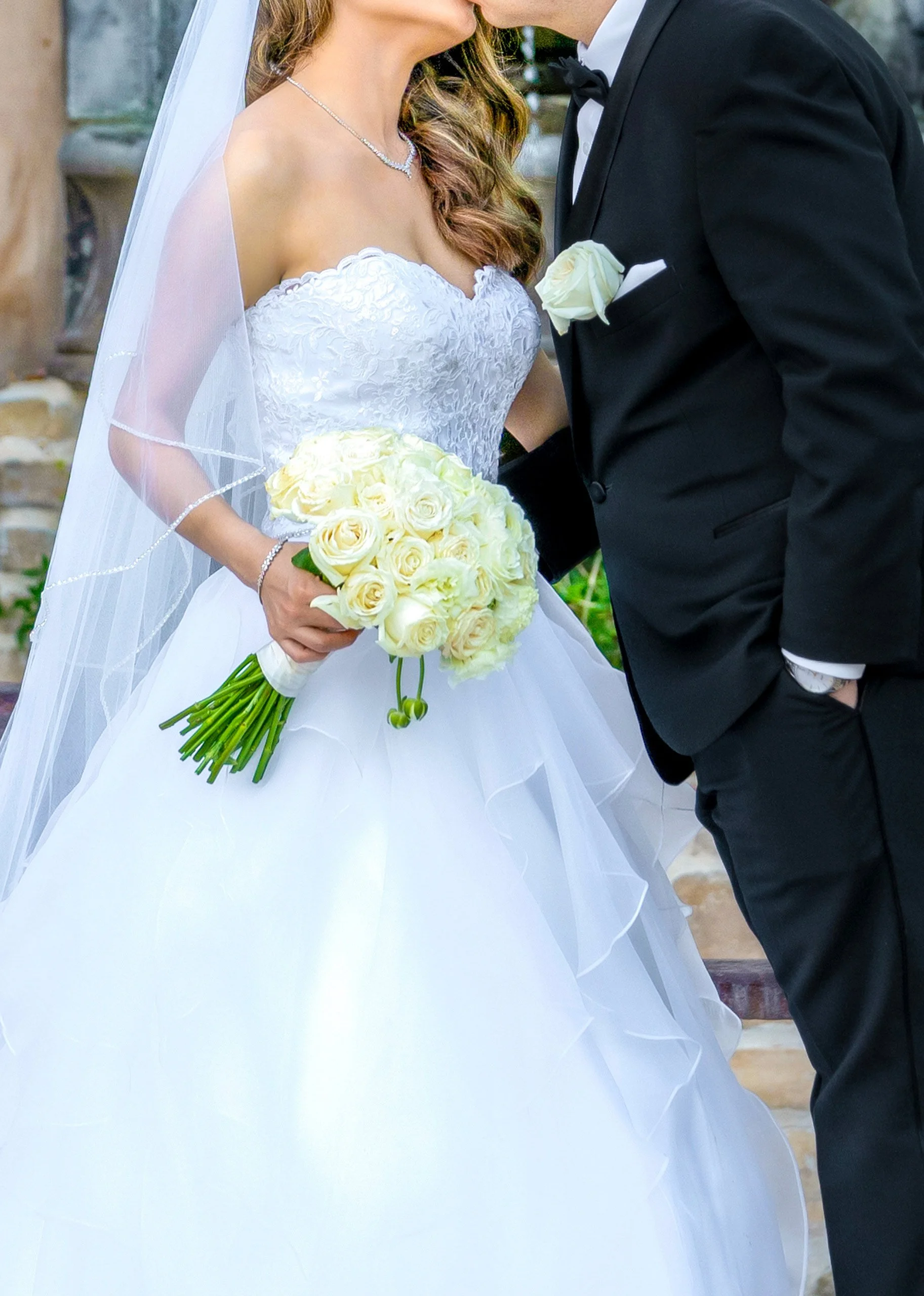 A bride in a white wedding dress holds a large bouquet of white roses while standing close to the groom in a black tuxedo.