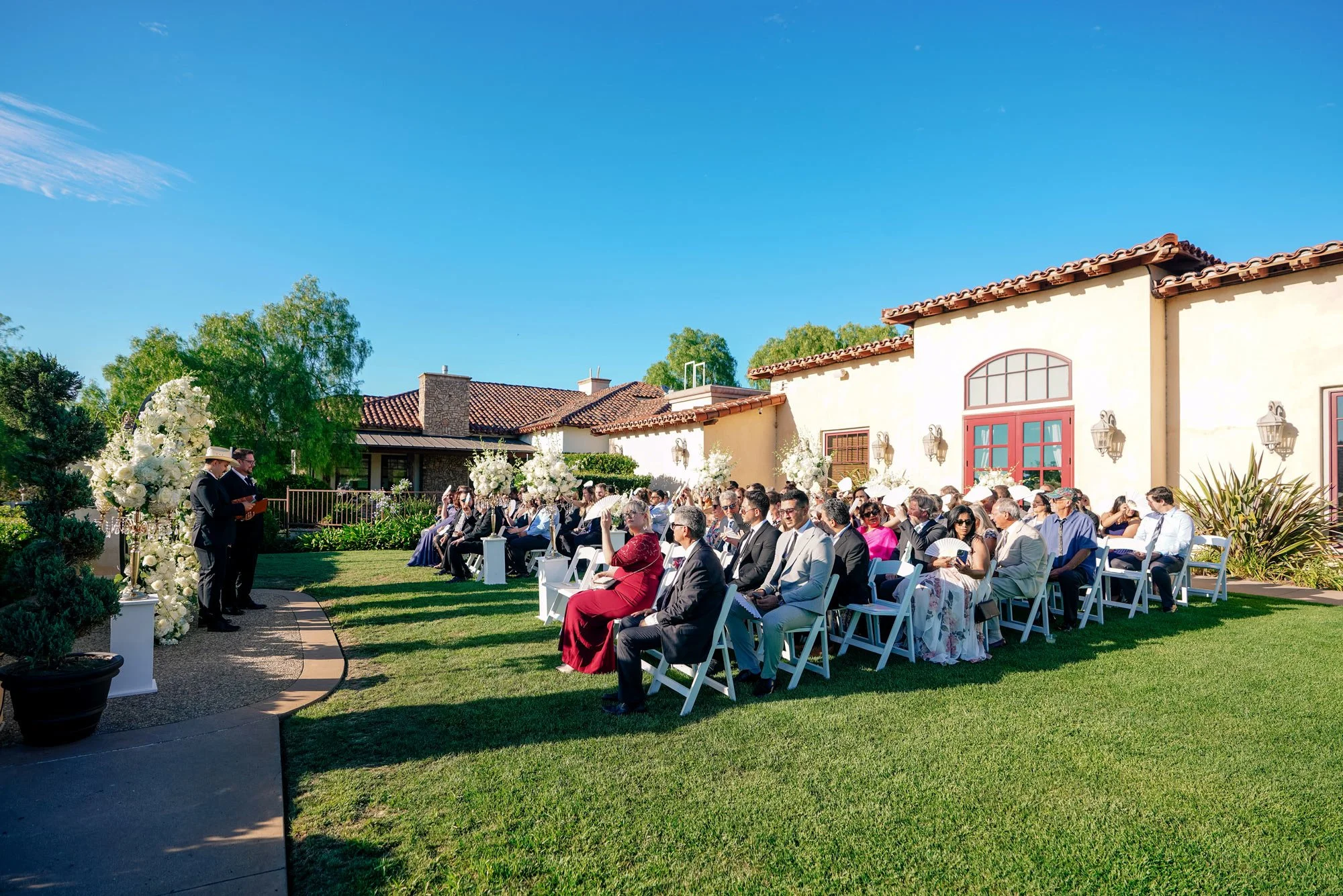 An outdoor photograph of a daytime wedding ceremony on a sunny day in Maderas Golf Club. Guests are seated in white folding chairs on a grassy lawn, watching as a man in a dark suit speaks at a podium at the front of the aisle. A large white floral a