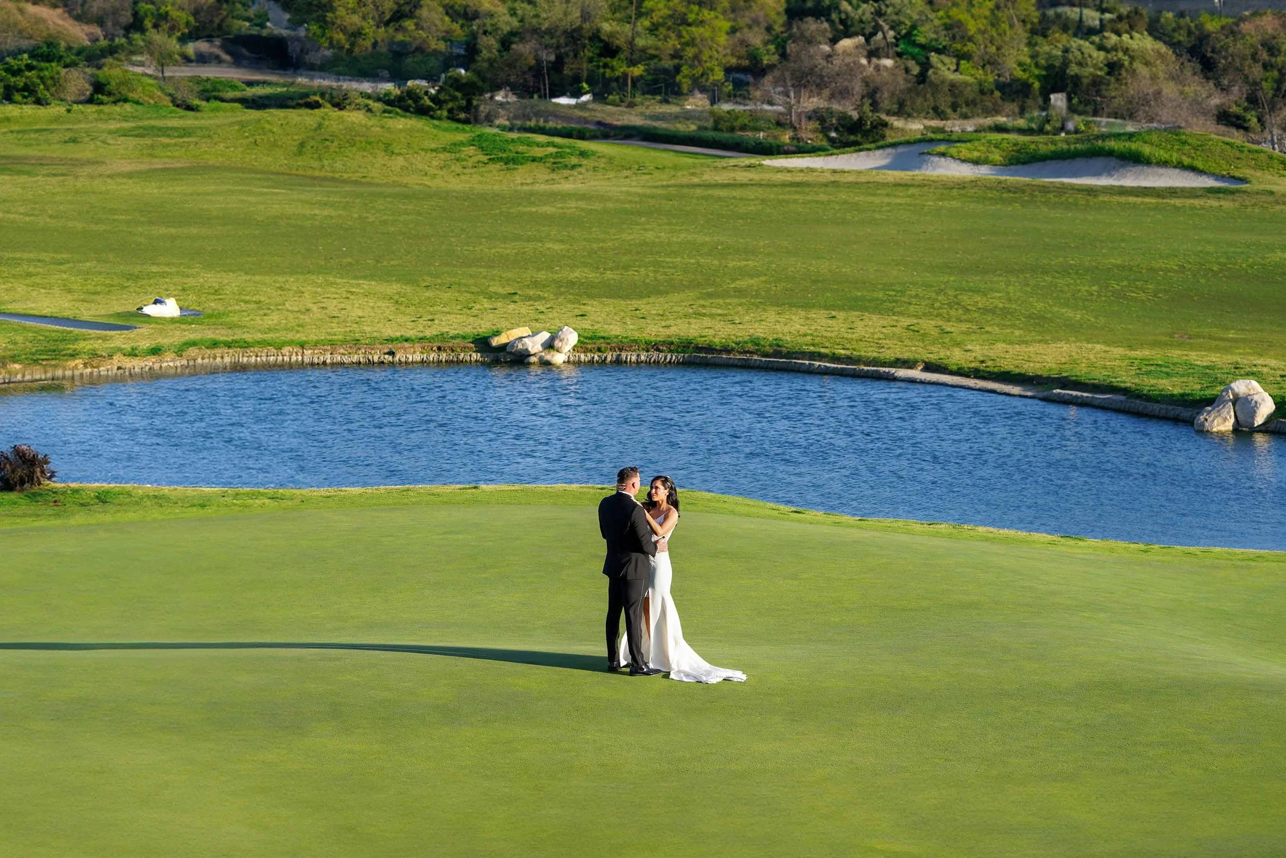 A full-length outdoor photograph of a newly married couple embracing on a grassy golf course. The groom wears a black tuxedo, and the bride wears a white wedding dress with a train. They are standing near a blue pond, with green fairways, sand bunker