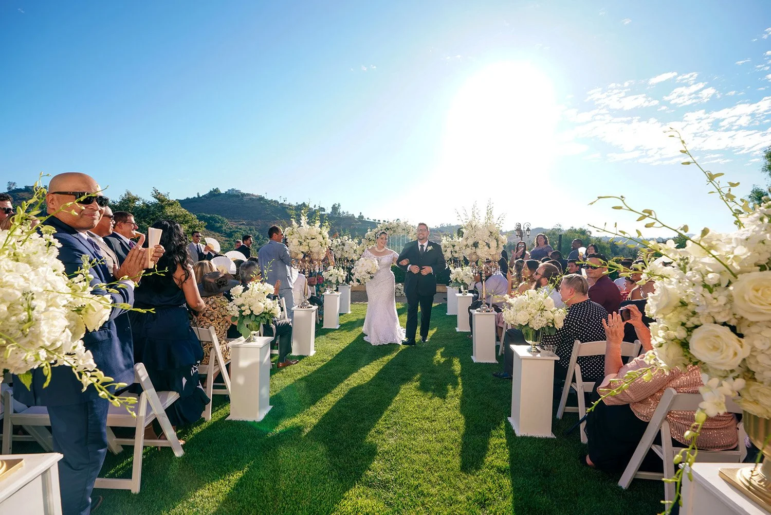 A bride and groom walk down a grassy aisle together after their outdoor wedding ceremony. They are surrounded by seated guests who are clapping and smiling. The aisle is lined with tall, white floral arrangements on pedestals, and the bright sun shin
