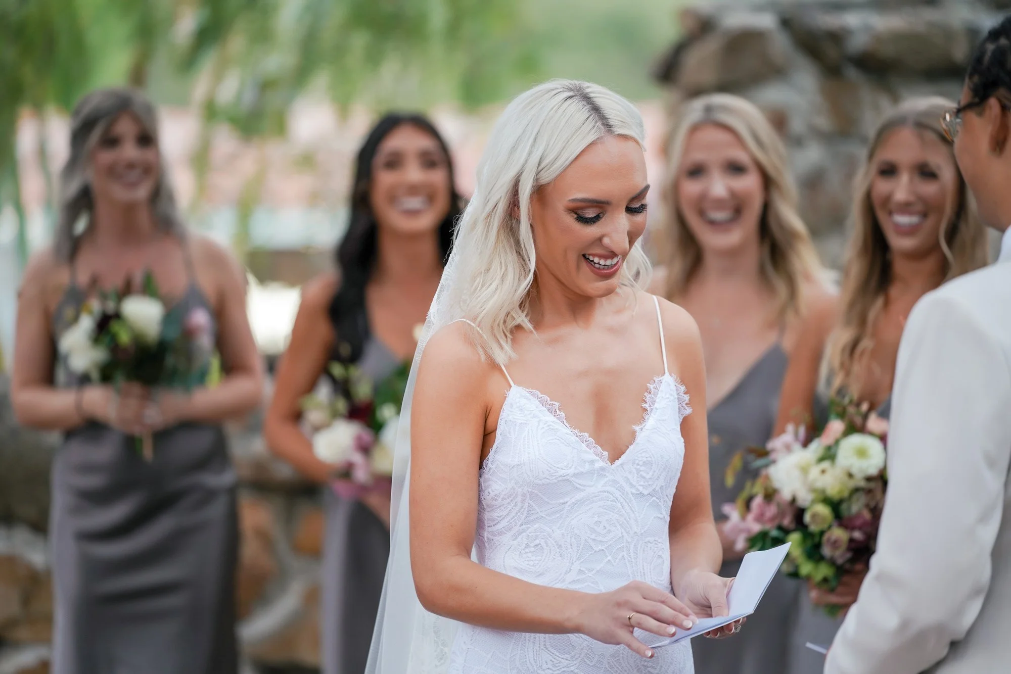 A photo of a bride in a white lace slip dress with spaghetti straps and a veil, looking down and smiling at a small white card she is holding, while three smiling bridesmaids in grey dresses stand behind her, and part of a groom in a cream-colored su