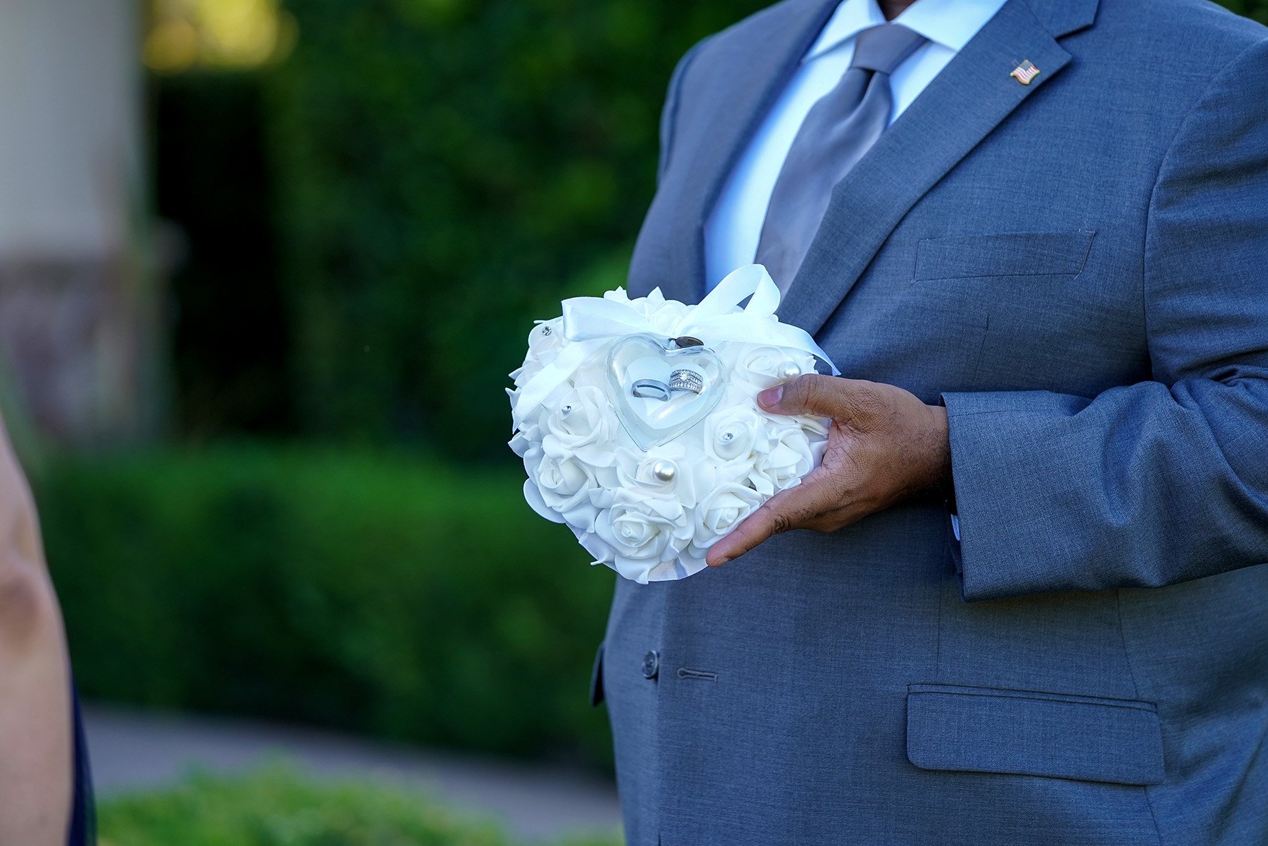 An individual in a grey suit holds a heart-shaped ring bearer pillow made of white fabric roses. A clear, heart-shaped container in the center of the pillow holds two wedding bands. The background shows a soft-focus view of lush green foliage and an 