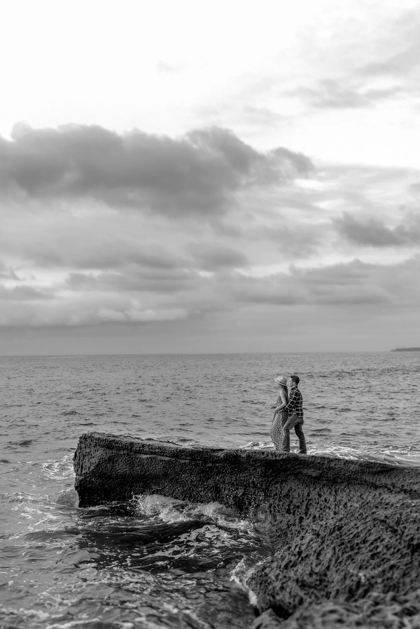 A black and white photo of a couple standing on a rock by the ocean, facing each other, with clouds in the sky.