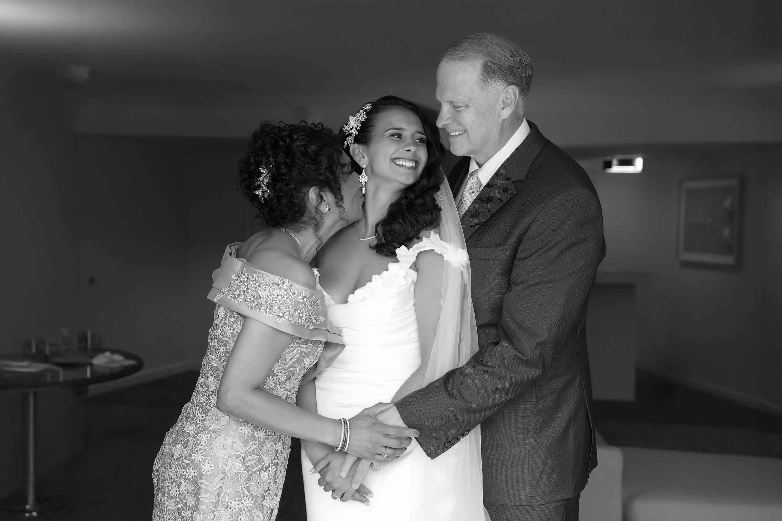 A tender black and white wedding portrait by Reimo Photography showing a bride smiling joyfully while being embraced by her parents. Her mother kisses her cheek while her father looks on with a smile, holding the bride's hands.