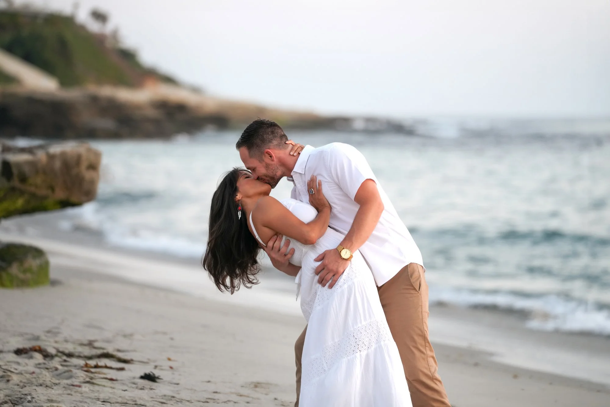 An outdoor photograph of a couple embracing and kissing on a sandy beach. The woman wears a white dress, and the man wears a white short-sleeved shirt and tan pants. Waves are rolling in on the shore, and a rocky bluff is visible in the background on