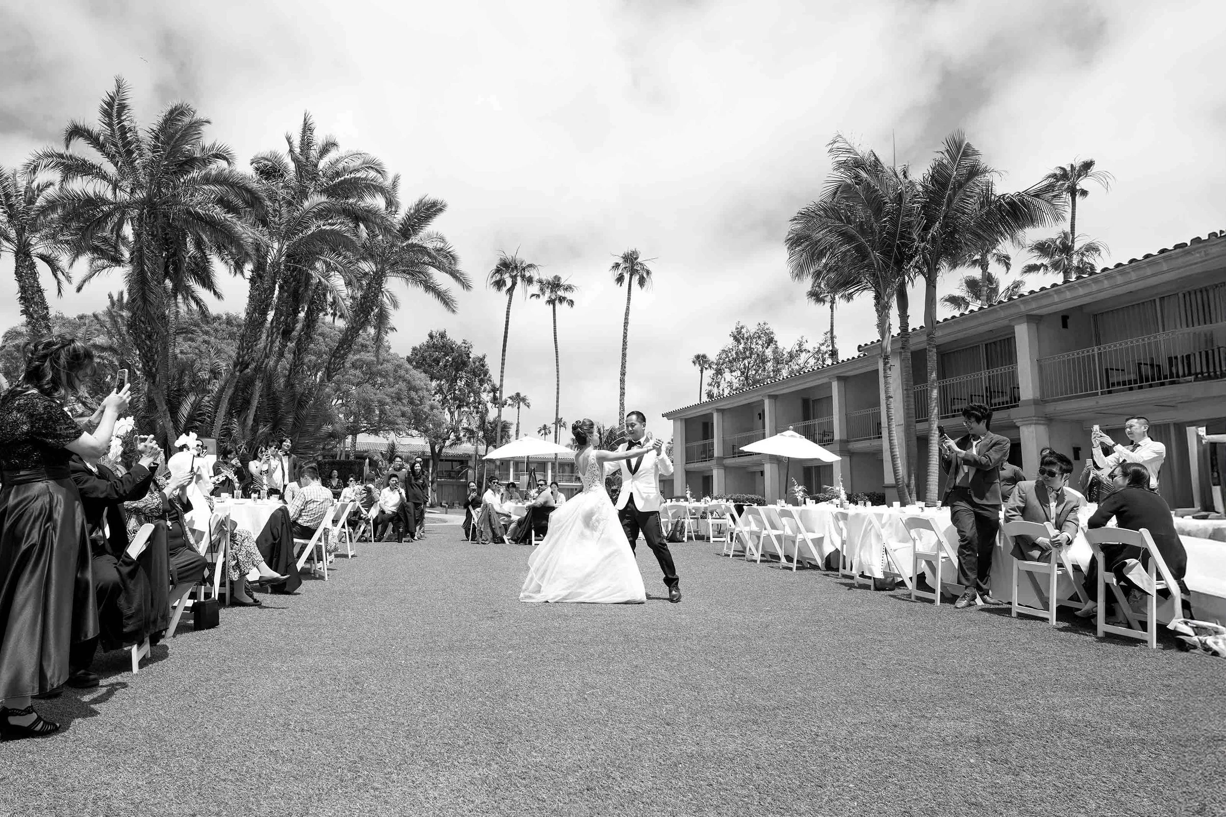 A black and white photograph captures a bride and groom sharing their first dance on a lawn at an outdoor wedding reception in San Diego, surrounded by guests, tall palm trees, and a resort building in the background.


