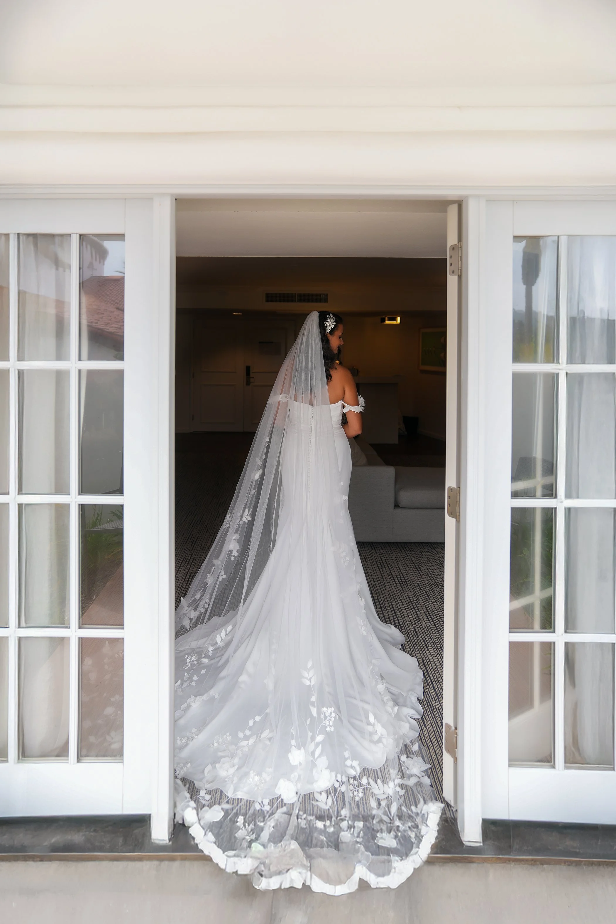 A full-length bridal portrait by Reimo Photography taken from behind. The bride, wearing a sleek white mermaid-style wedding gown, stands in an open set of white French doors at Omni La Costa Golf Club in Carlsbad. Her long, cathedral-length veil, ad