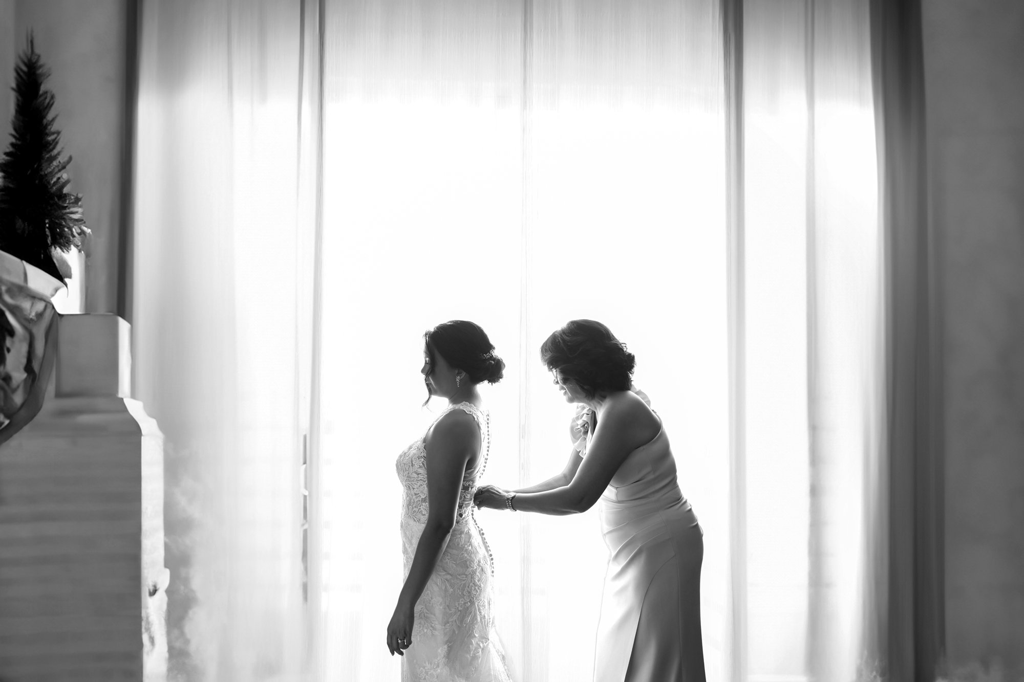 A black and white photo of a woman helping a bride button the back of her wedding dress in front of a bright window in San Diego.