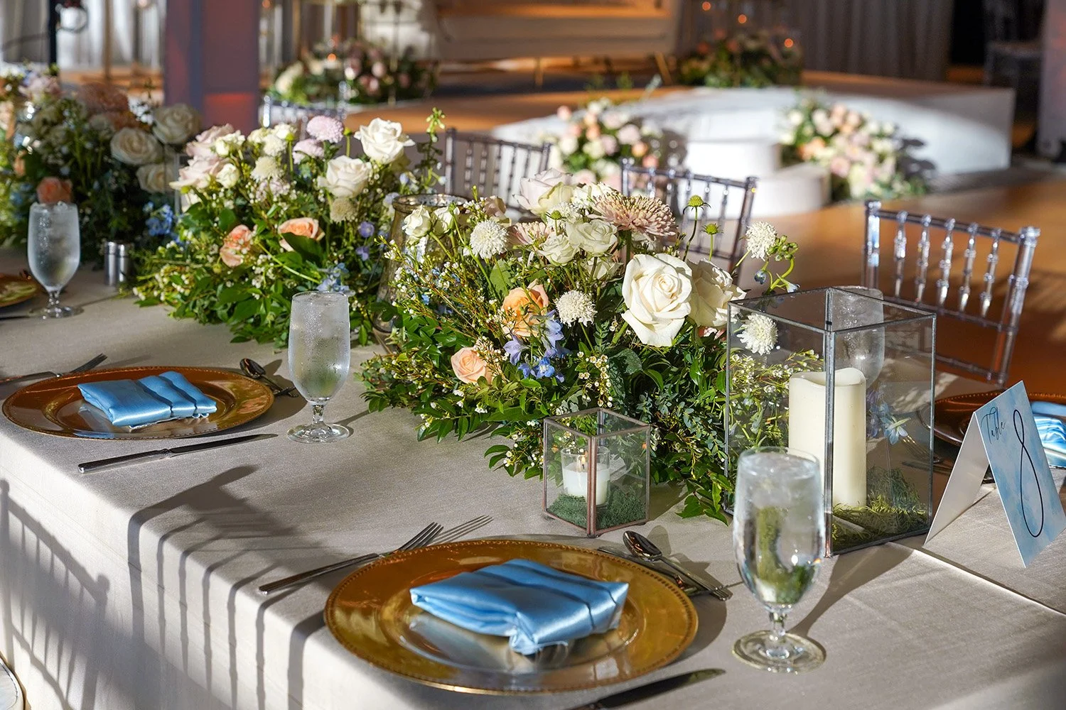 A formal dining table set for an event, featuring gold charger plates, light blue napkins, lush white and peach floral centerpieces, glass candle lanterns, and clear event chairs.
Would
