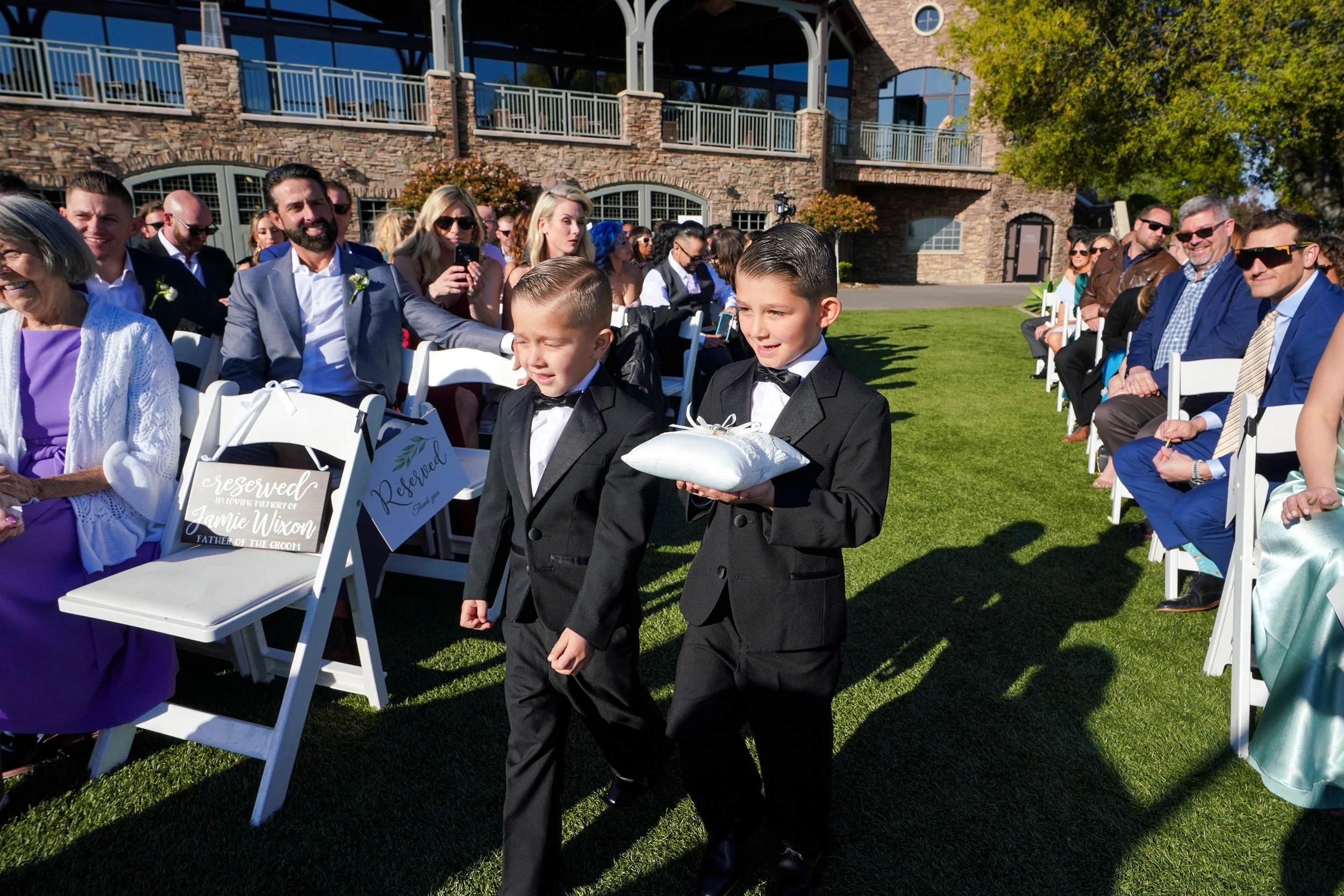 The ceremony is set on a lush lawn with guests seated in white folding chairs on either side. In the background, a large, two-story stone and wood building with a balcony overlooks the event under a clear blue sky. A reserved sign on the front-row ch