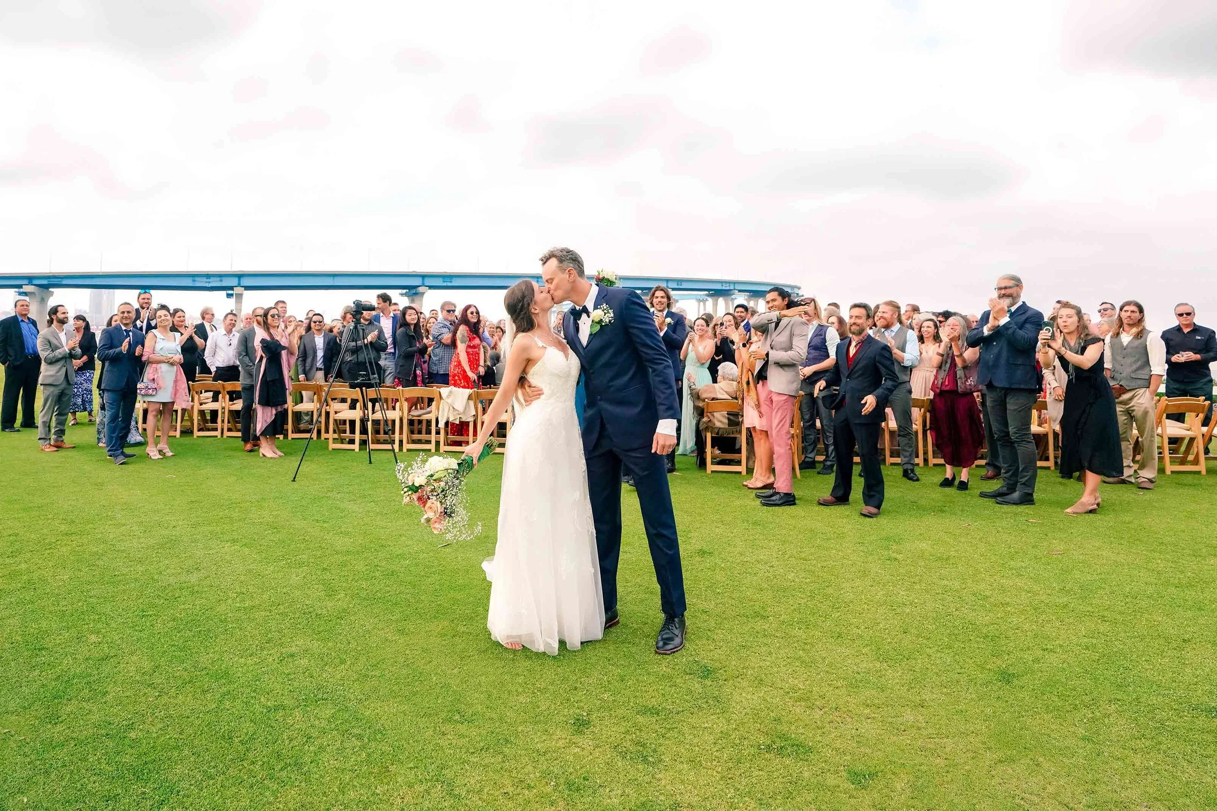 A bride in a white dress and a groom in a navy suit share a kiss in the foreground on a large green lawn, while wedding guests stand and applaud in the background under a cloudy sky.