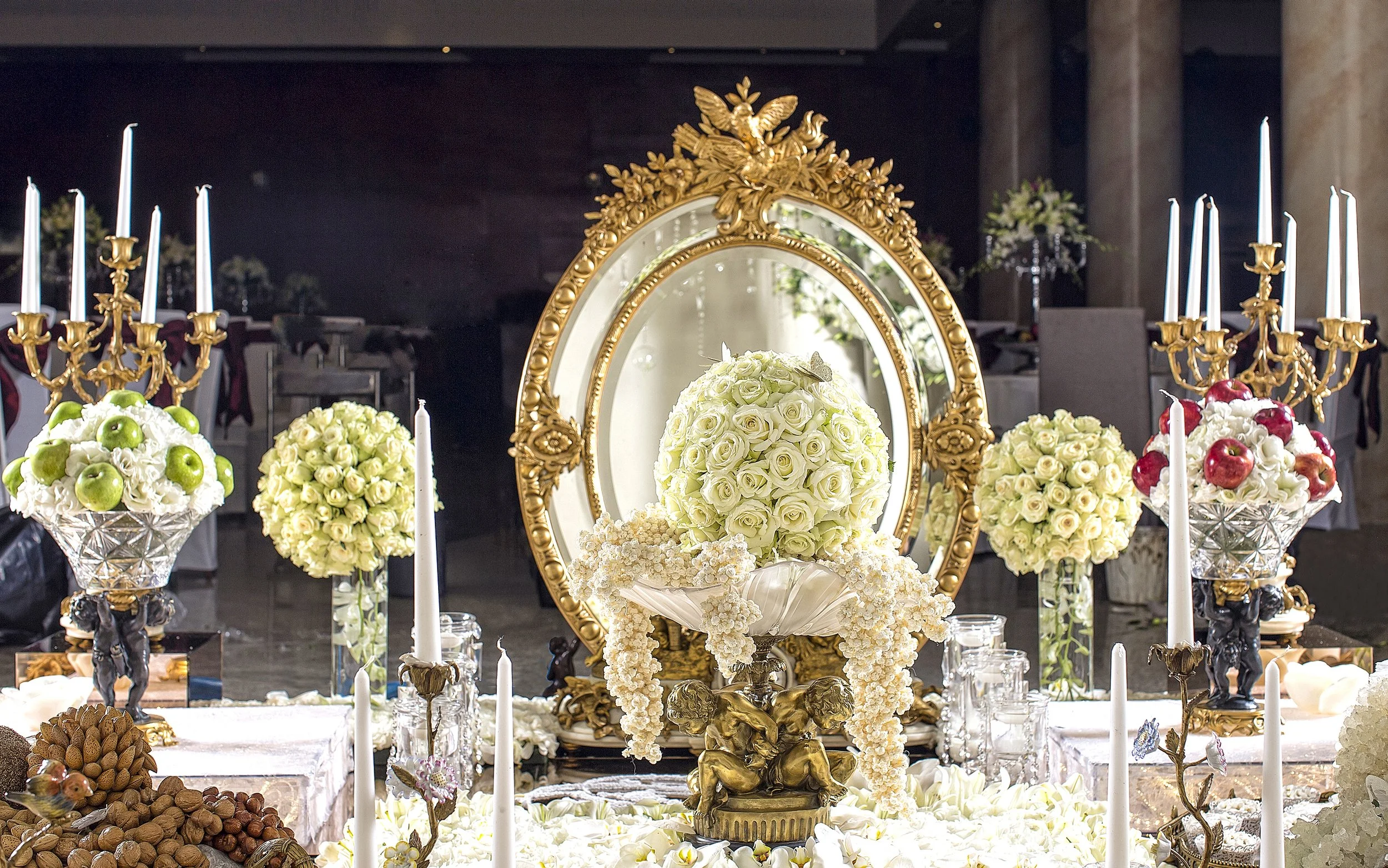 An elaborate, traditional Persian wedding table setting (Sofreh Aghd) featuring a large ornate gold mirror, candelabras with white taper candles, spheres of white roses and green apples, and various symbolic items like nuts and fruits on a white tabl