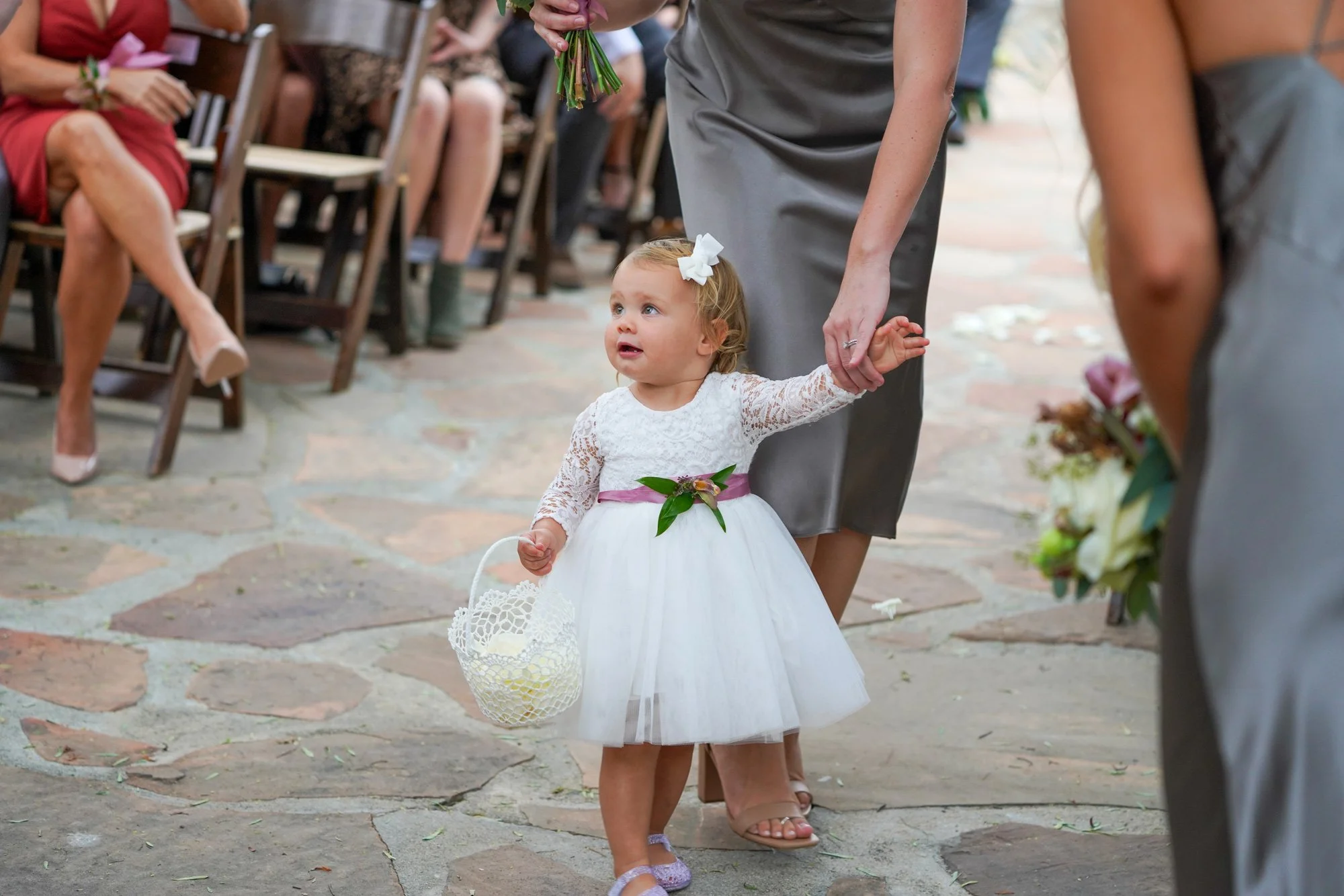 A photo of a young flower girl wearing a white lace dress with a full tulle skirt and a white bow in her hair, walking down a flagstone path while holding a white basket, guided by a person in a grey dress.




