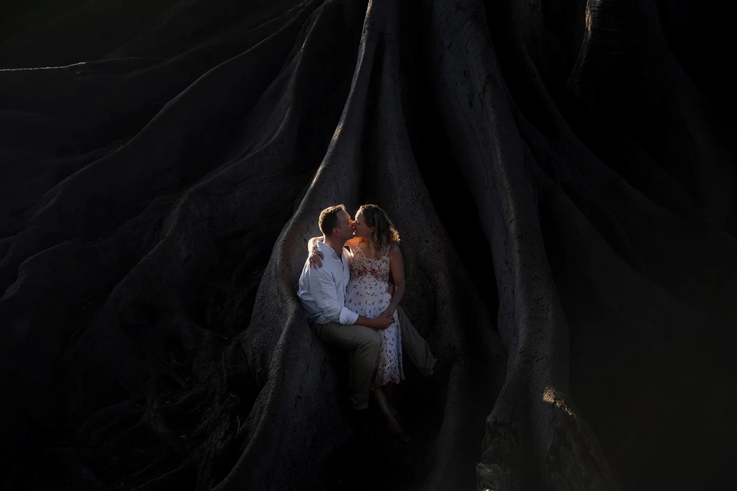 A couple sitting within the large, dark, exposed roots of a massive tree, sharing a kiss. The man wears a white long-sleeved shirt and tan pants, and the woman wears a light-colored floral dress. The lighting highlights the couple and the deep recess