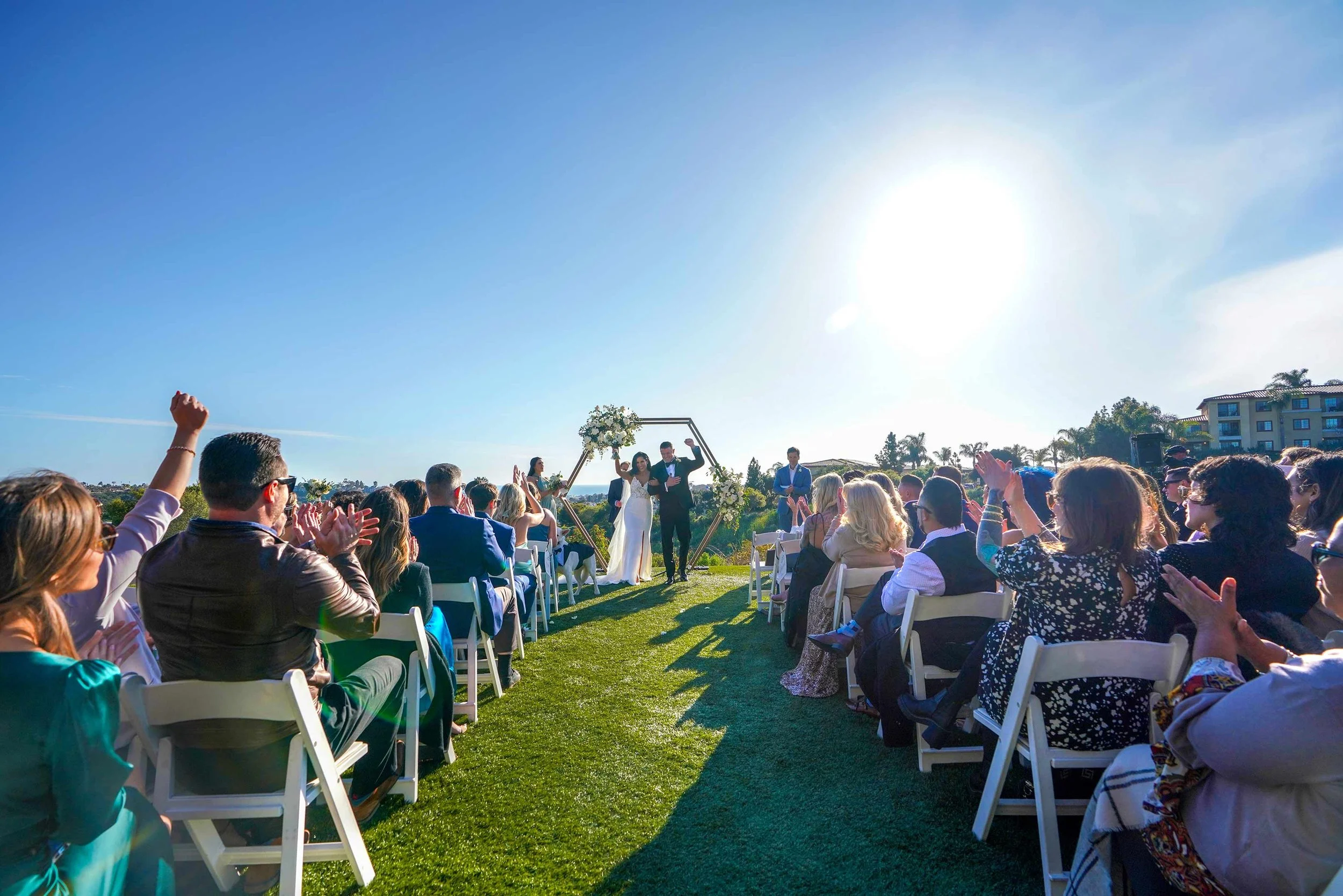 The couple stands under a geometric archway adorned with flowers, surrounded by seated guests in white chairs, all on a bright, sunny day with views of the golf course and hills in the background. The primary purpose of alt text is accessibility for 
