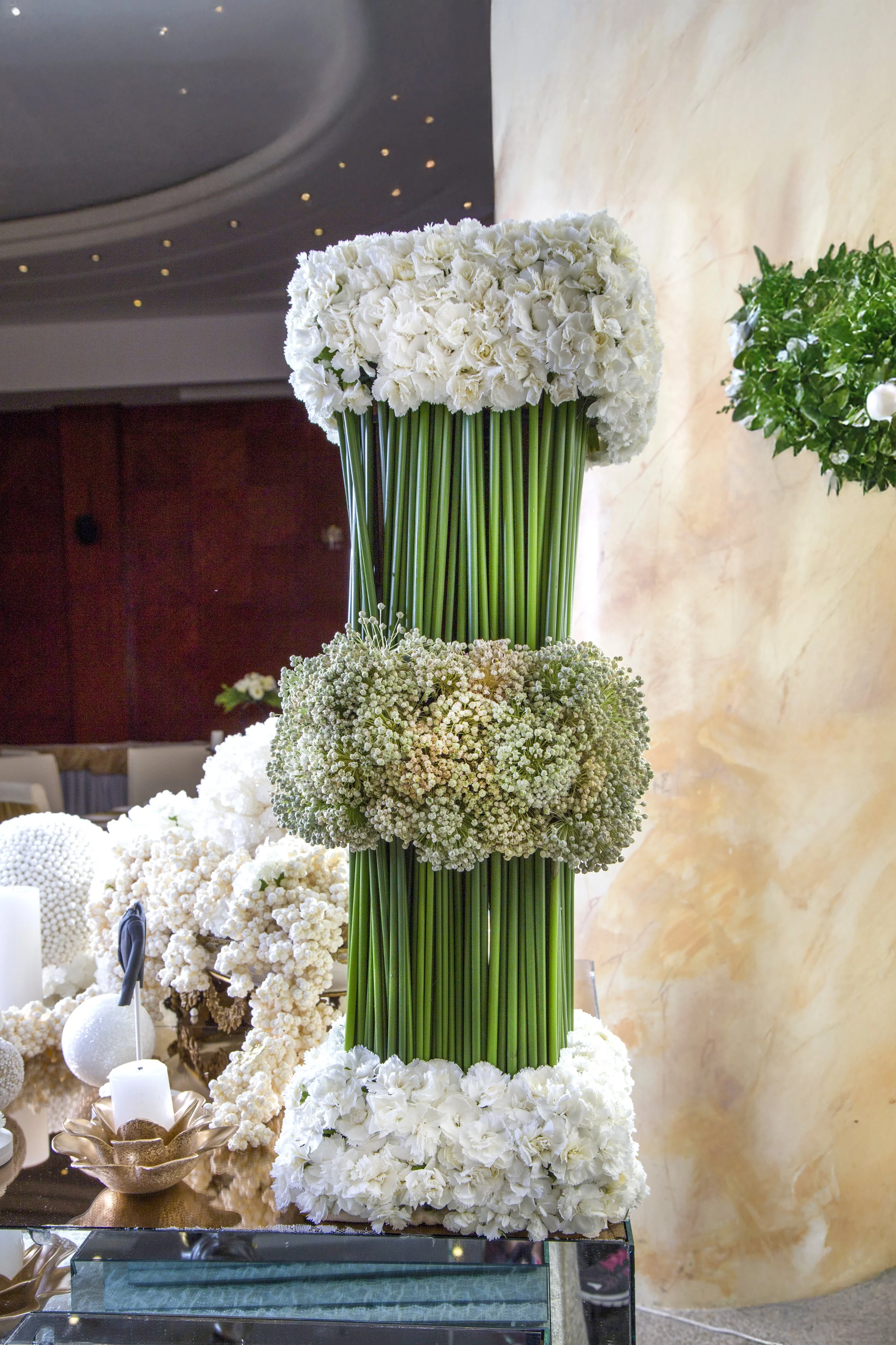 A tall, modern floral arrangement featuring vertical green stems flanked by bands of white and light green flowers, sitting on a mirrored table with additional white decor and candles.
