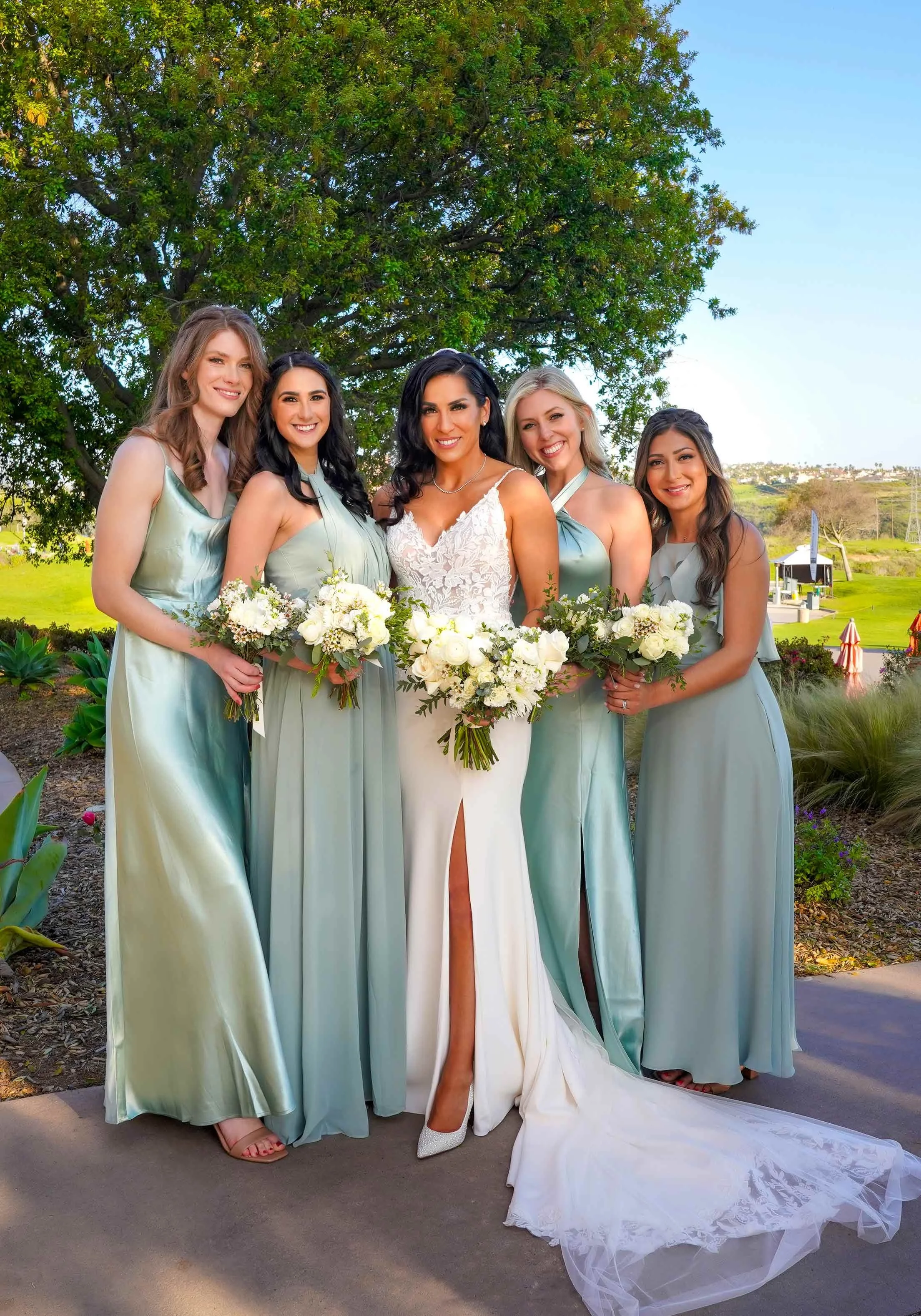 An outdoor photograph of a bride and four bridesmaids posing together on a paved area with green landscaping in the background. The bride stands in the center wearing a white lace wedding dress with a high leg slit and a long train. The four bridesma