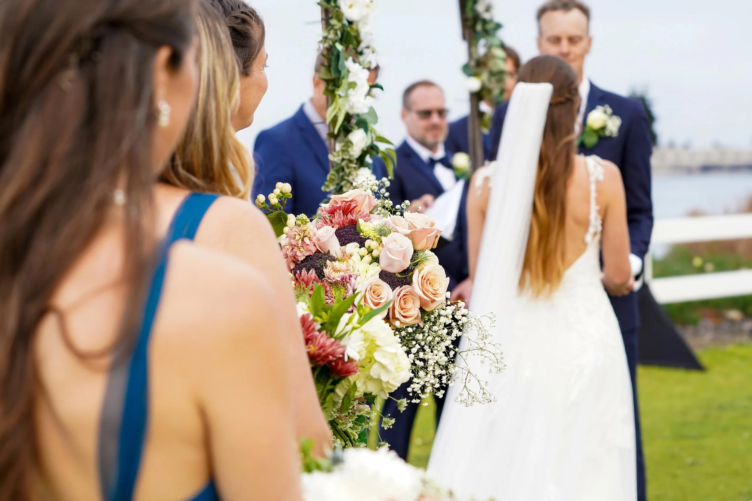 A close-up shot from behind the bridesmaids at a wedding ceremony, focusing on a large bouquet of pink, peach, and white flowers with the bride and groom standing at the altar in the background.


