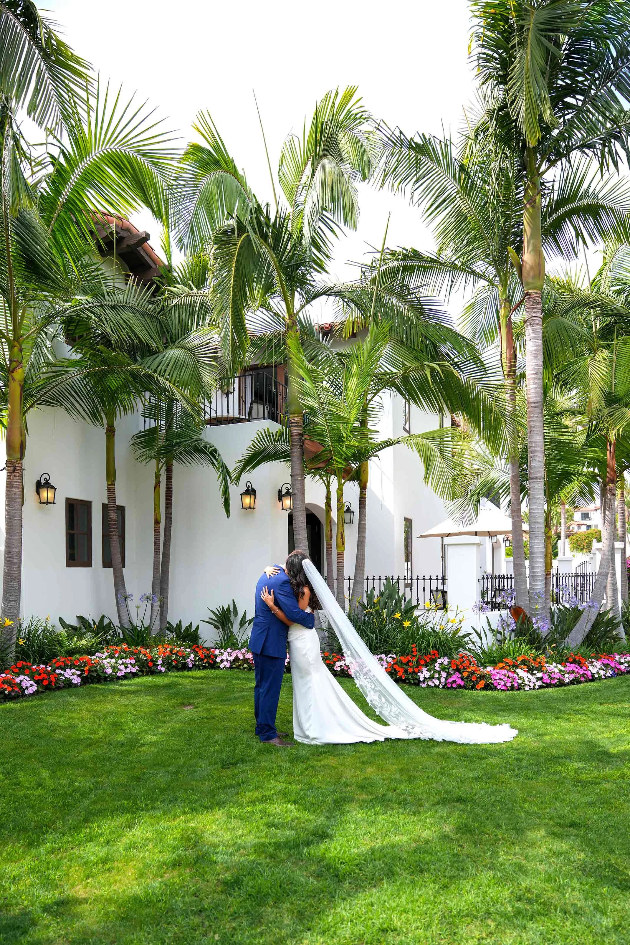 A couple share a kiss on a green lawn, standing in front of a white Spanish-style building surrounded by tall palm trees and colorful flowers.