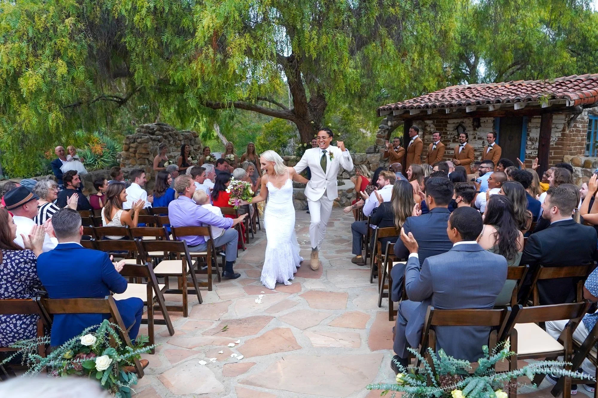 A newly married couple smiles and raises their hands in celebration as they walk down the stone aisle after their outdoor wedding ceremony at the rustic Leo Carrillo Ranch Historic Park.