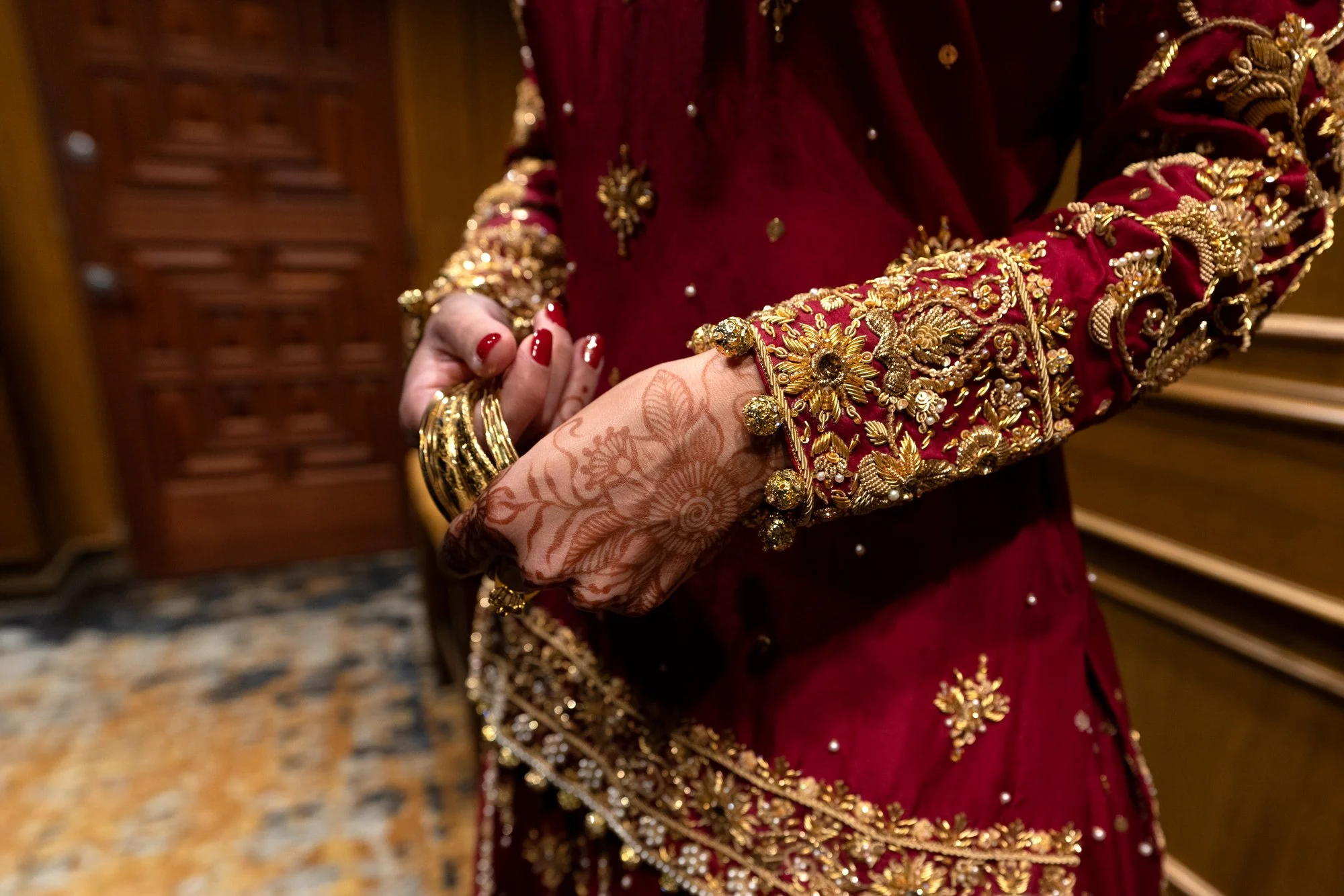 A close-up photograph of a person's hands and arms, adorned with henna, gold jewelry, and a vibrant red and gold traditional garment. The person is holding a stack of gold bangles in their left hand, which has red-painted fingernails. The garment fea