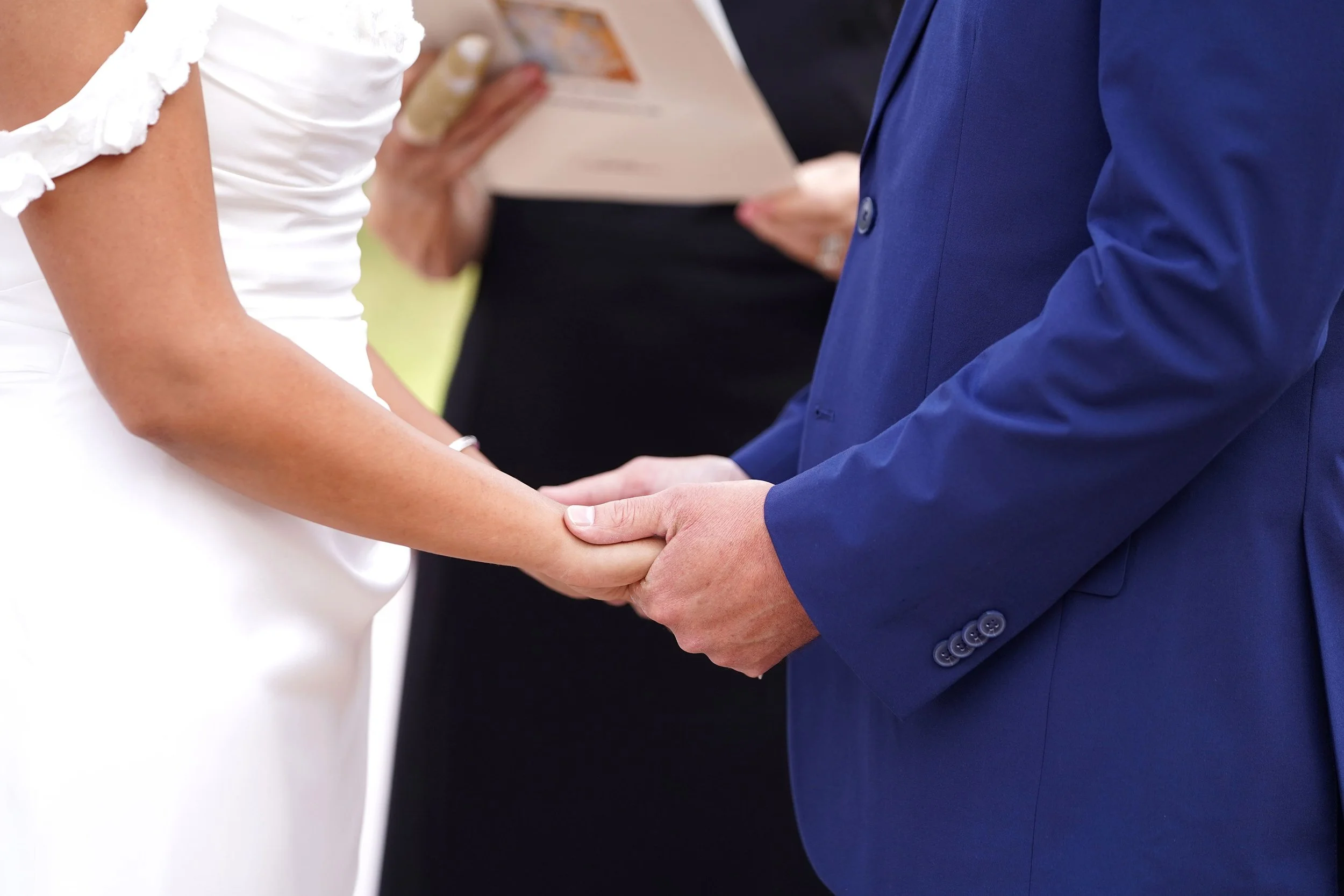 A close-up photograph of a bride in a white dress and a groom in a blue suit holding hands during their wedding ceremony.


