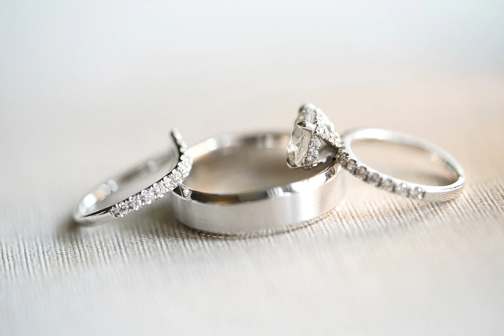 A close-up photograph of three wedding rings, including a thick smooth band, a diamond engagement ring, and a thinner diamond-studded band, resting on a light-colored, textured surface.


