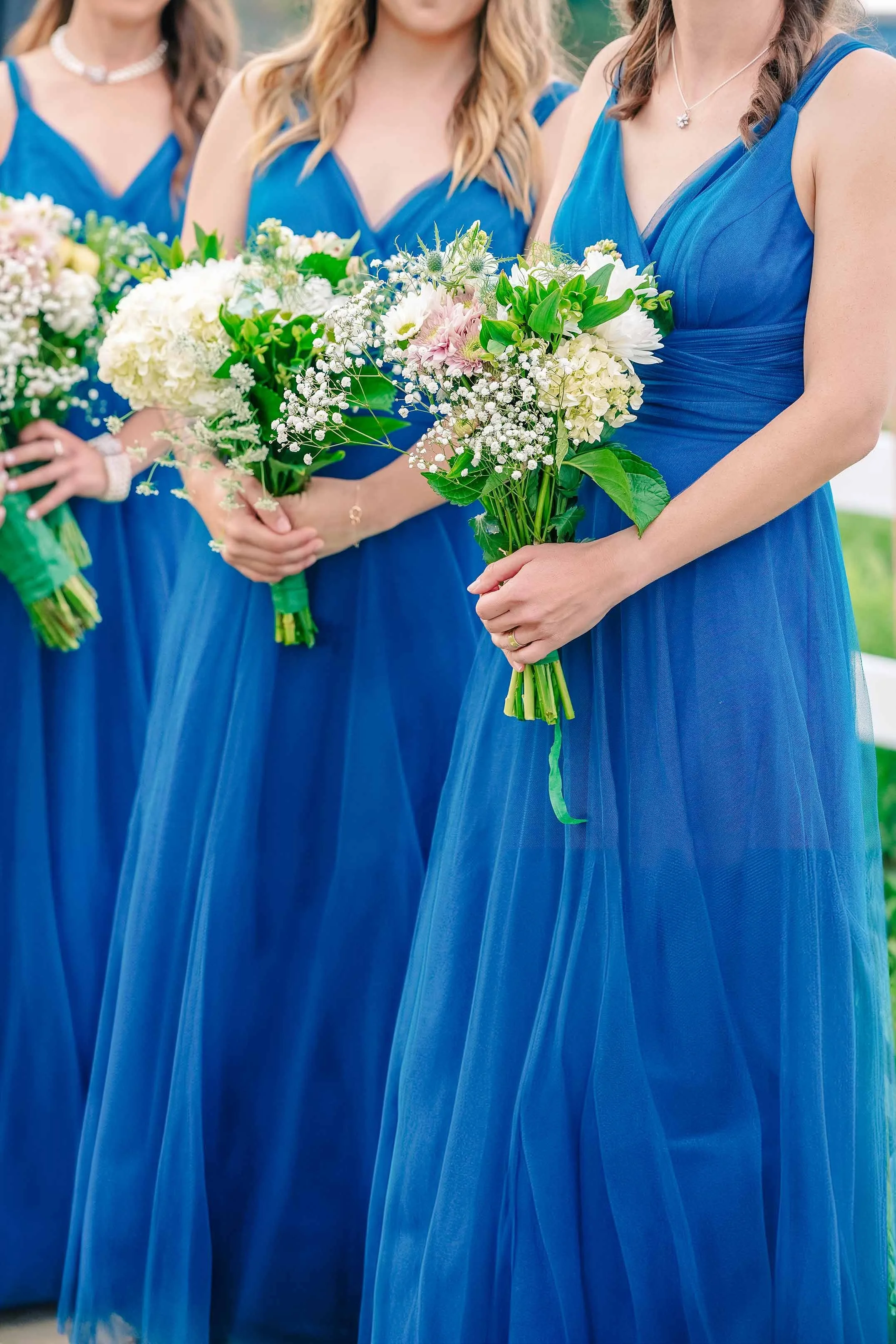 Three bridesmaids standing side-by-side, wearing matching royal blue dresses and holding bouquets of white, green, and pink flowers.


