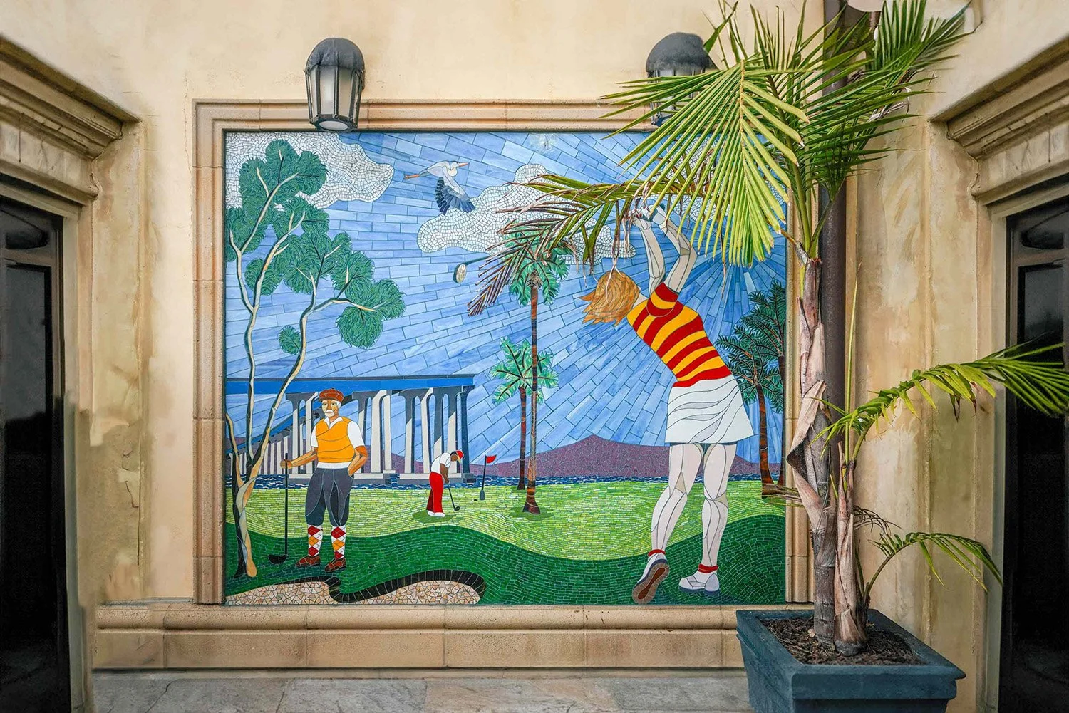 A vibrant outdoor mosaic artwork depicting two golfers on a green course, one in an orange vest leaning on a club and the other in a red shirt bending to putt, with a classical building, palm trees, and a blue sky in the background in Coronado.