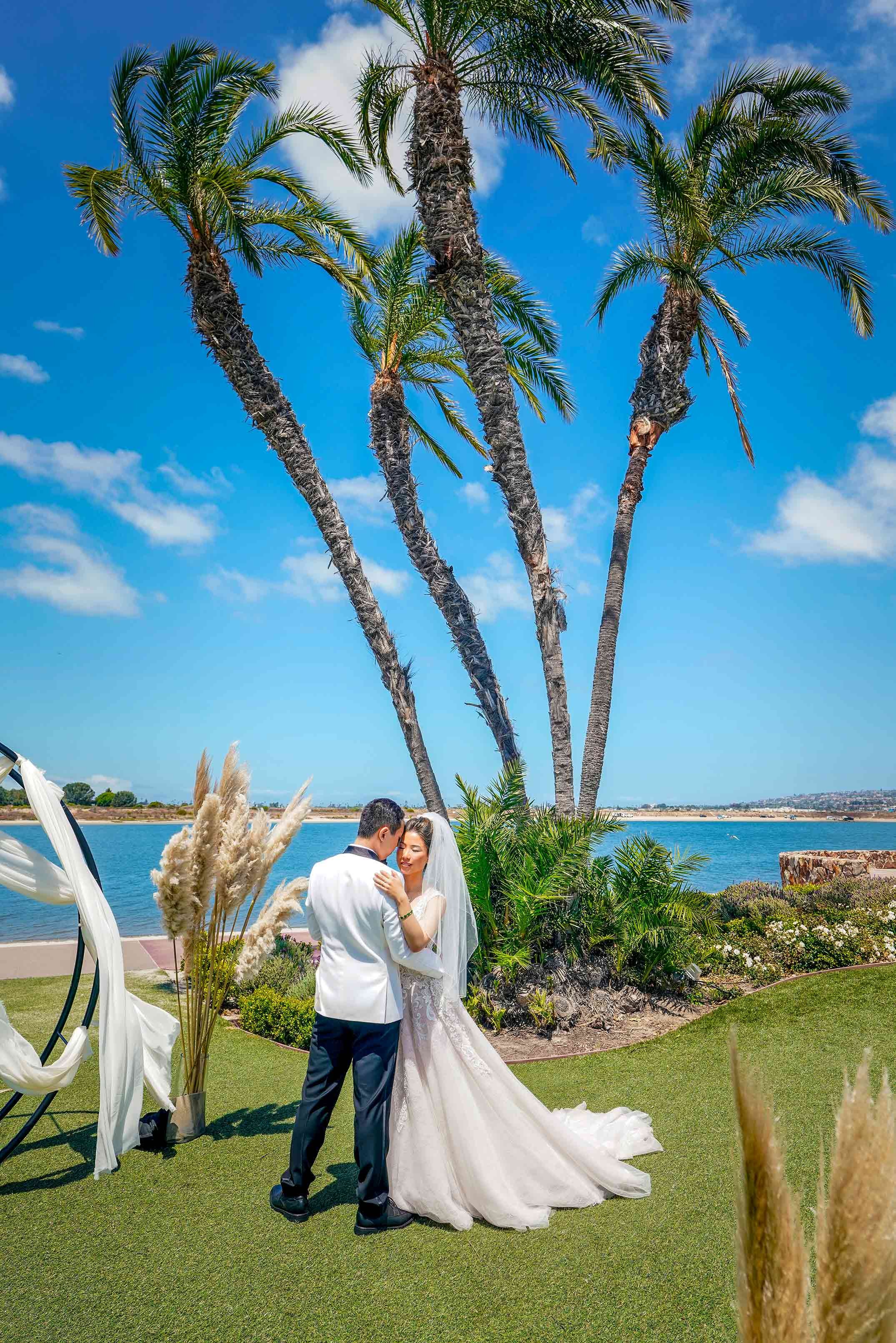 A bride in a lace wedding gown and a groom in a white tuxedo jacket share a moment embracing under tall palm trees, overlooking the blue water during their sunny outdoor wedding in Mission Bay, San Diego


