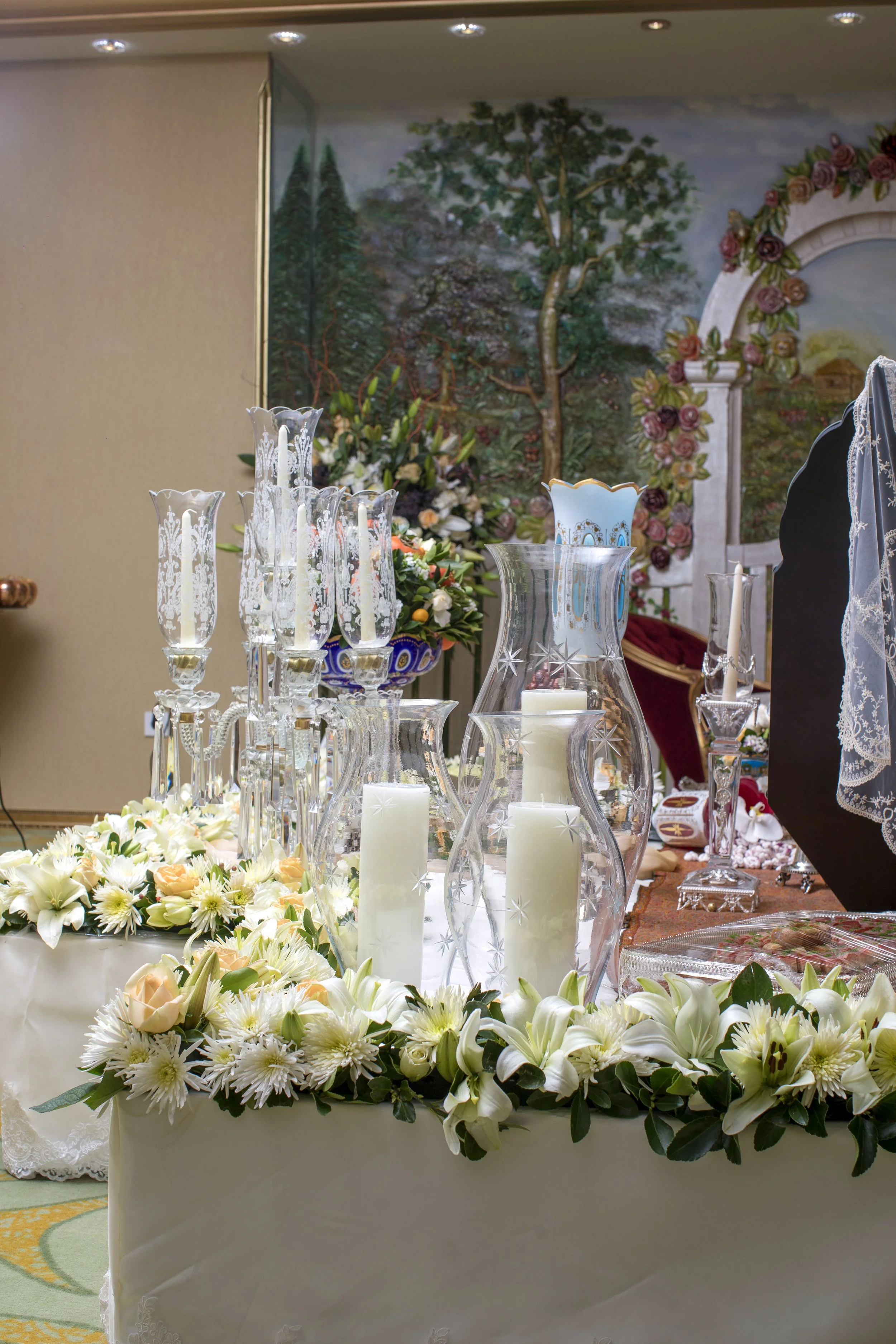 An elaborate, formal table setting for a Persian wedding ceremony (Sofreh Aghd) featuring tall crystal candelabras and glass vases with white pillar candles and fresh white floral arrangements. The display is set against a backdrop of a painted mural