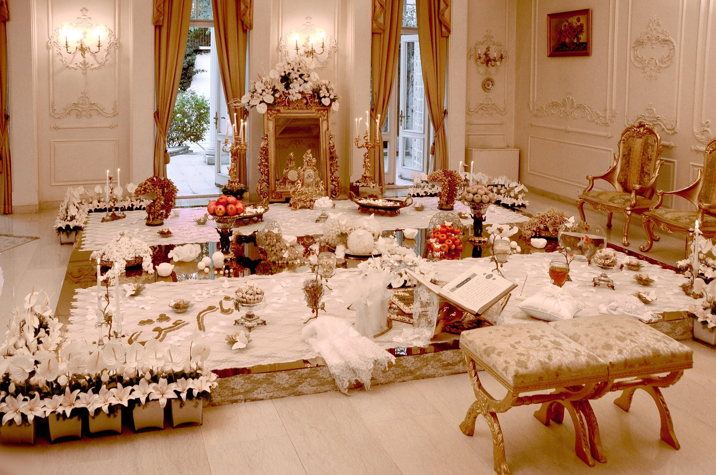An elaborate, traditional Persian wedding table setting (Sofreh Aghd) in an elegant room, featuring a large ornate gold mirror, candelabras, a holy book, fruits, nuts, and extensive white floral and gold decorations.
