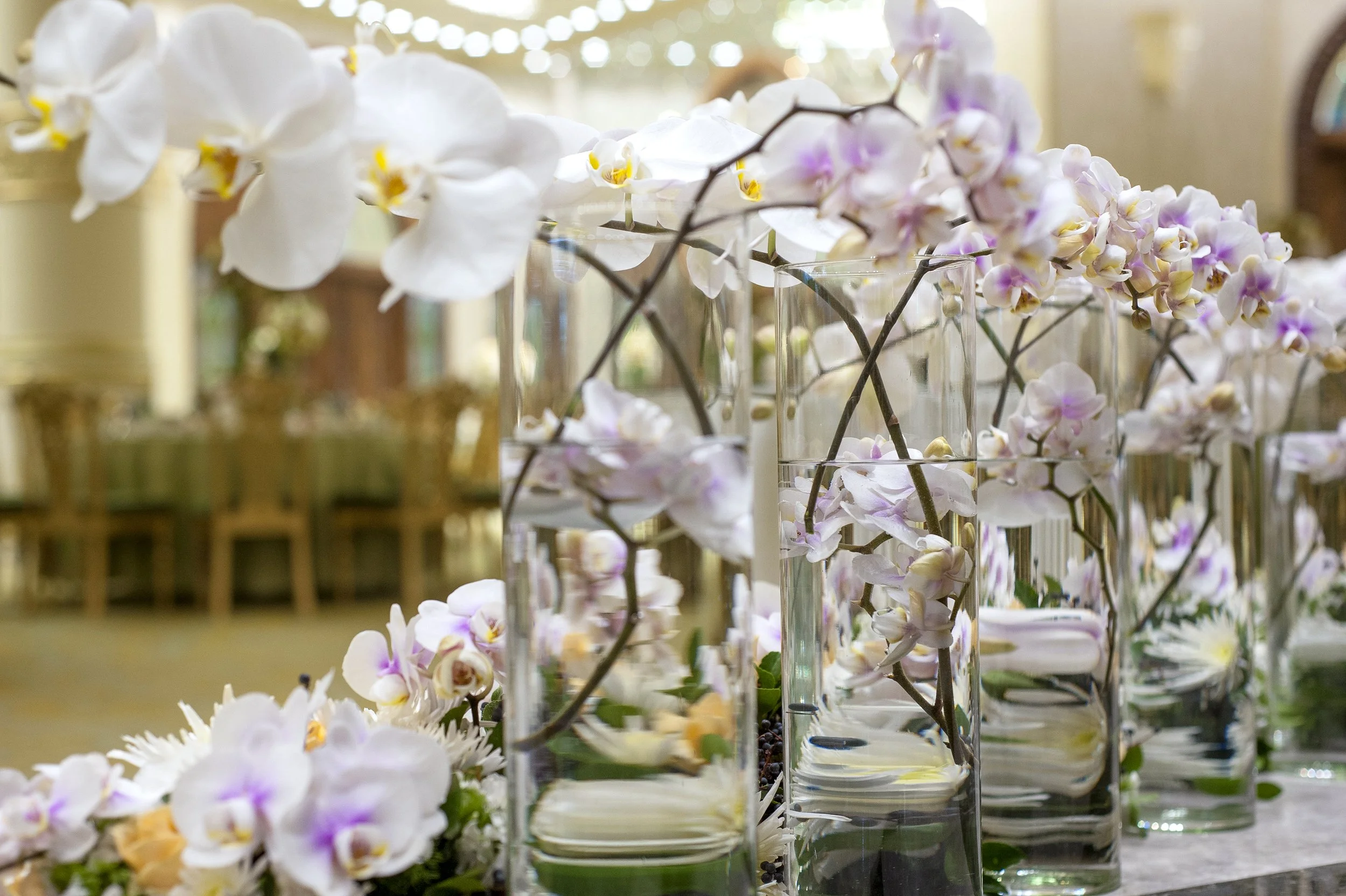 A long row of elegant, modern table centerpieces featuring white and light pink orchid stems submerged in tall, clear glass vases filled with water, set up for a formal event in a banquet hall.

