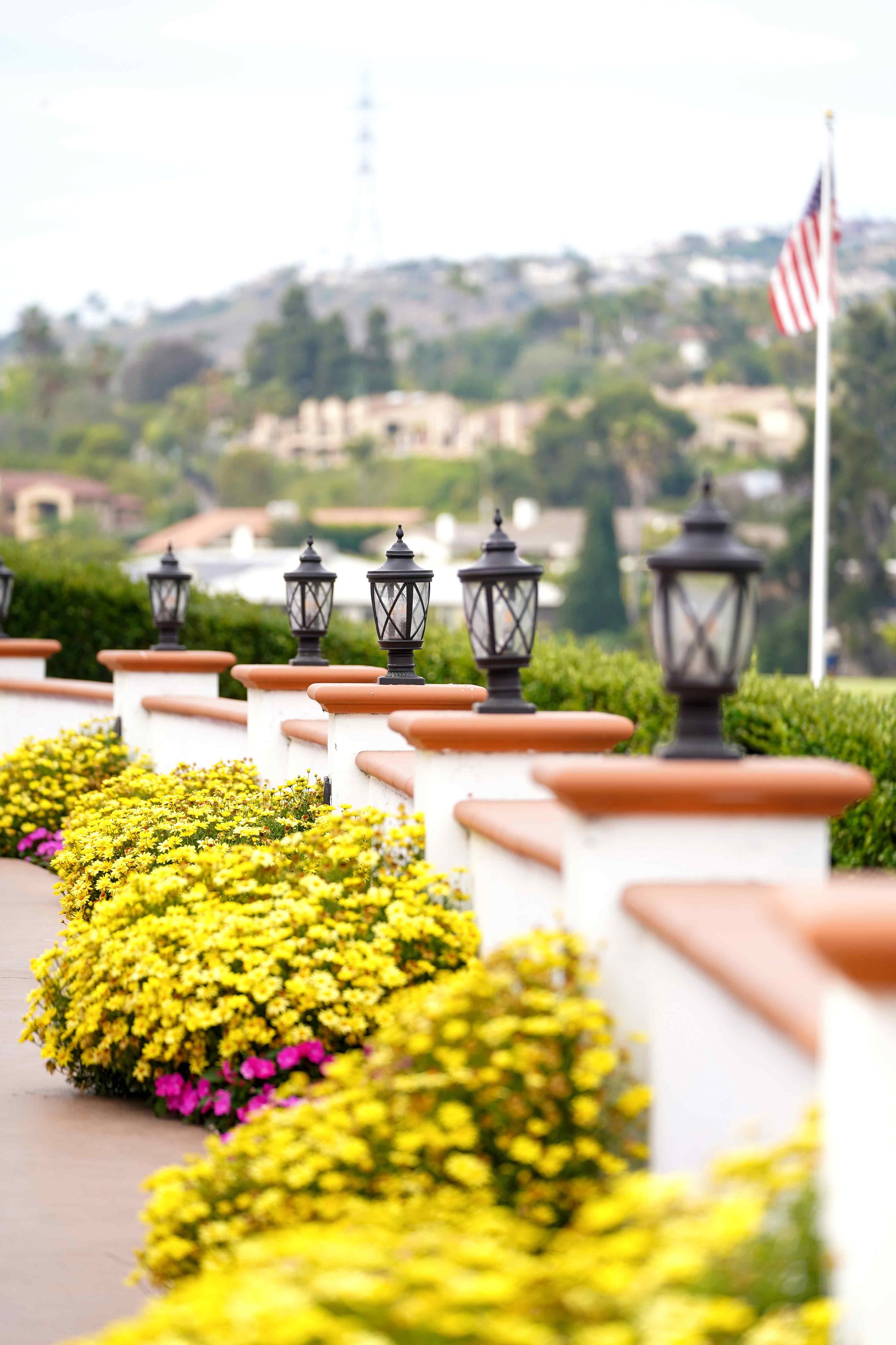 A white wall with terracotta caps and black post lamps lined with bright yellow and pink flowers leads diagonally into the distance, with a blurred background of hills and an American flag at Omni Resort in La Costa.