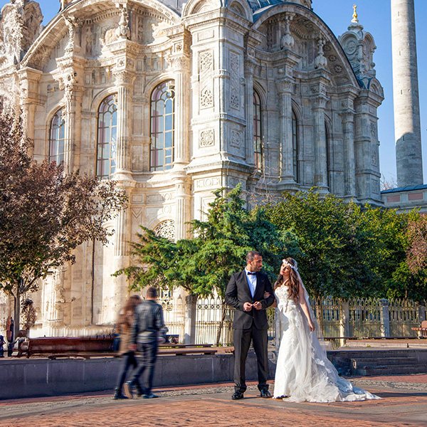 A bride and groom stand together in an outdoor courtyard in front of the grand Ortaköy Mosque in Istanbul. The ornate, Baroque-style mosque features intricate marble carvings and tall arched windows. 