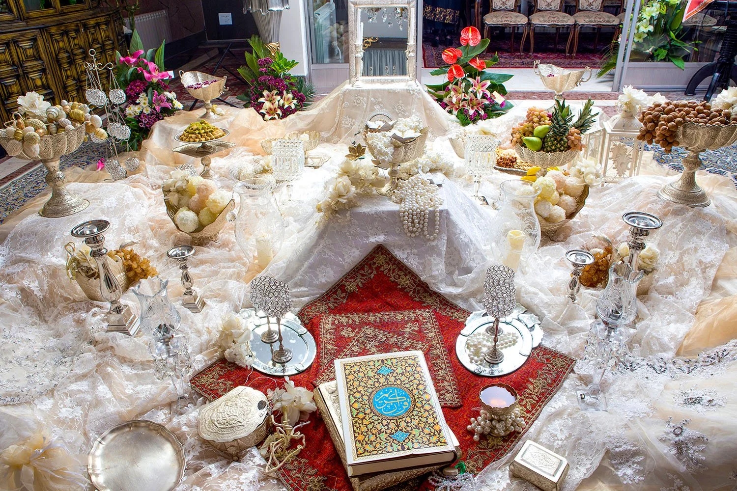 An elaborate, traditional Persian wedding table setting (Sofreh Aghd) featuring an ornate mirror, candelabras, a holy book, nuts, sweets, and flowers on a white lace tablecloth