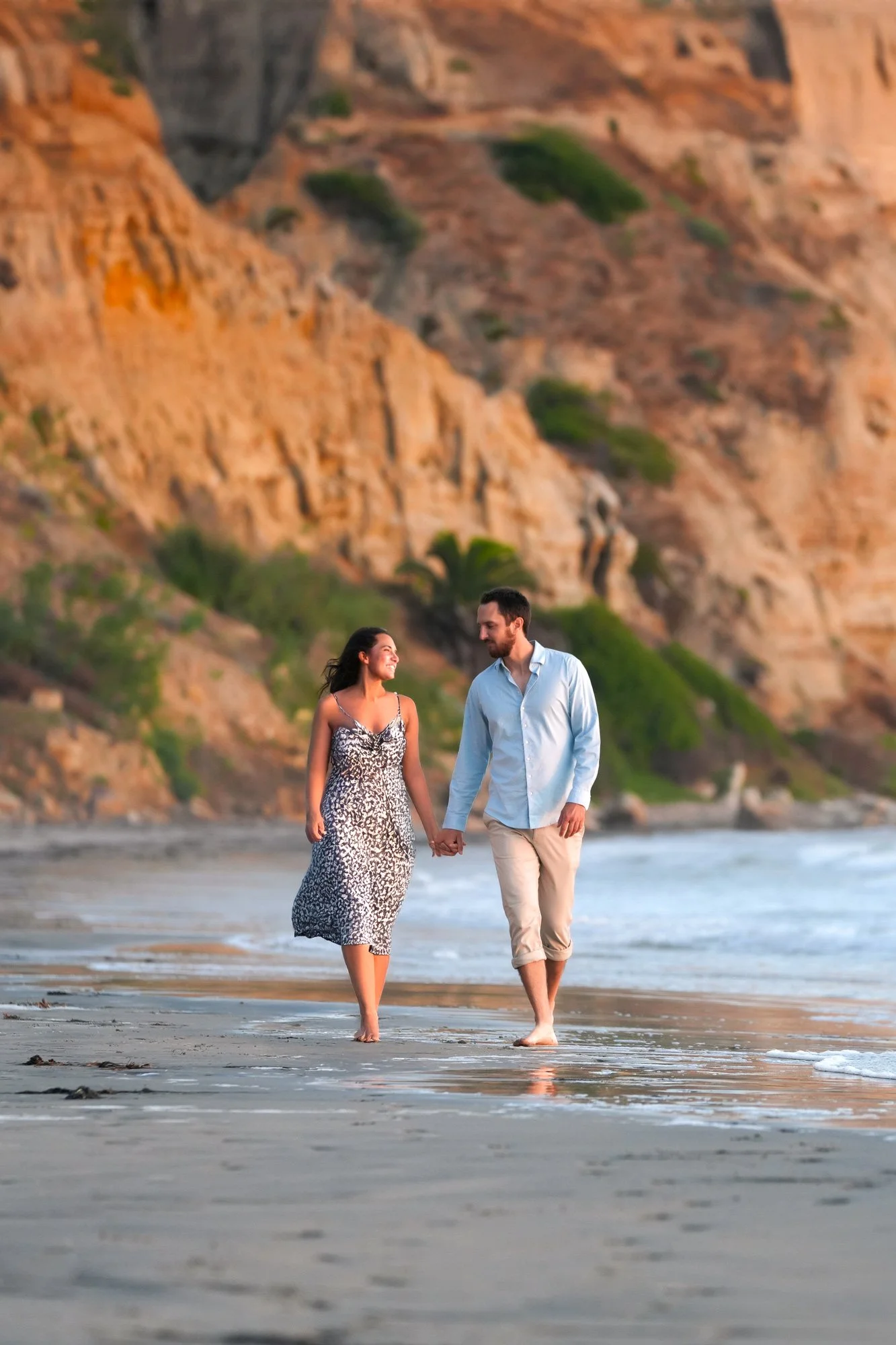 A couple walks hand-in-hand along the beach at sunset, with rocky cliffs and greenery in the background.