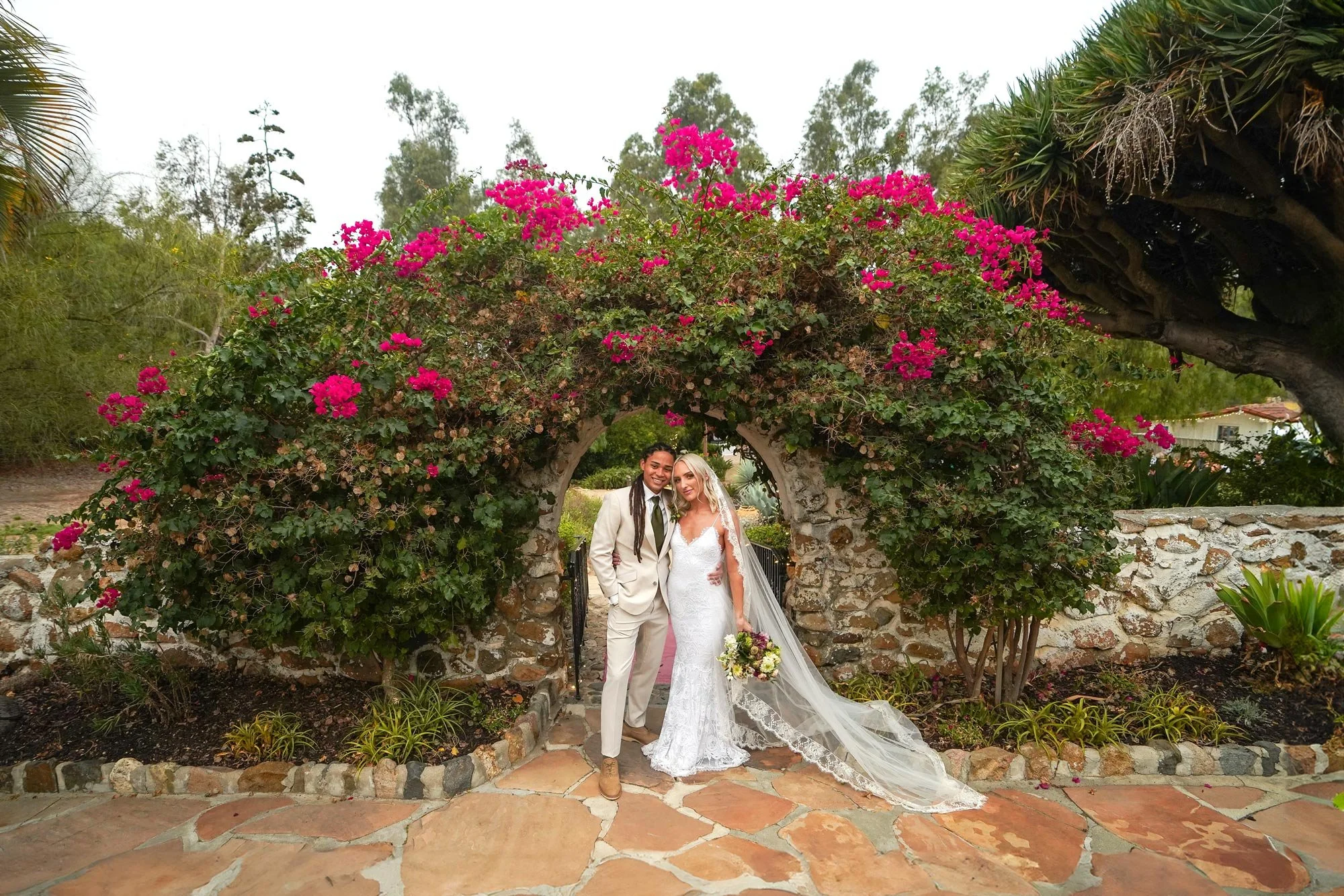 A photo of a newly married couple posing for a portrait under an archway overflowing with bright pink bougainvillea flowers, with one wearing a cream-colored suit and the other in a white lace wedding dress with a long veil trailing on the flagstone 