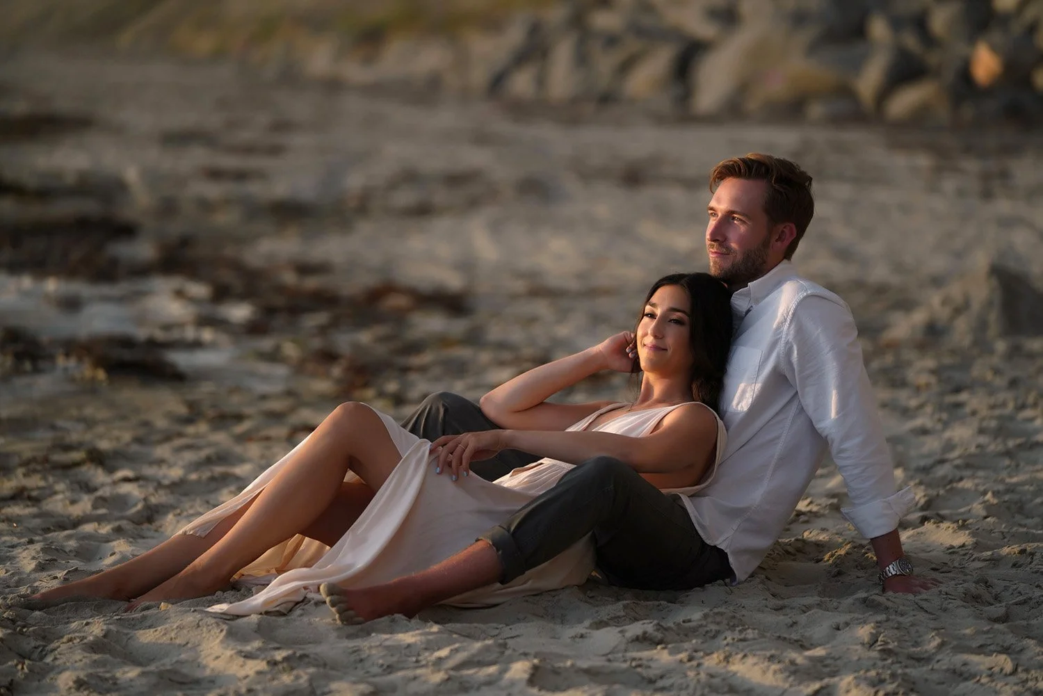 A couple sitting on a sandy beach during sunset or sunrise. The woman, in a light-colored dress, leans against the man, who wears a white long-sleeved shirt and dark pants and is looking out towards the light. They are sitting close together in the s