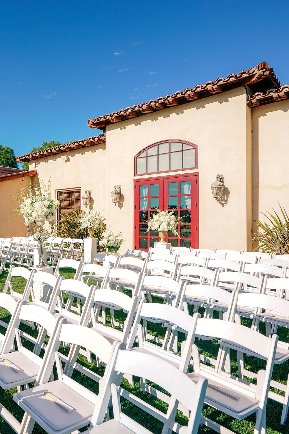 An empty wedding ceremony setup on a grassy lawn. Numerous rows of white folding chairs face a Spanish-style stucco building with a red-tiled roof and vibrant red double doors at Maderas Golf Club. Large white floral arrangements are placed near the 