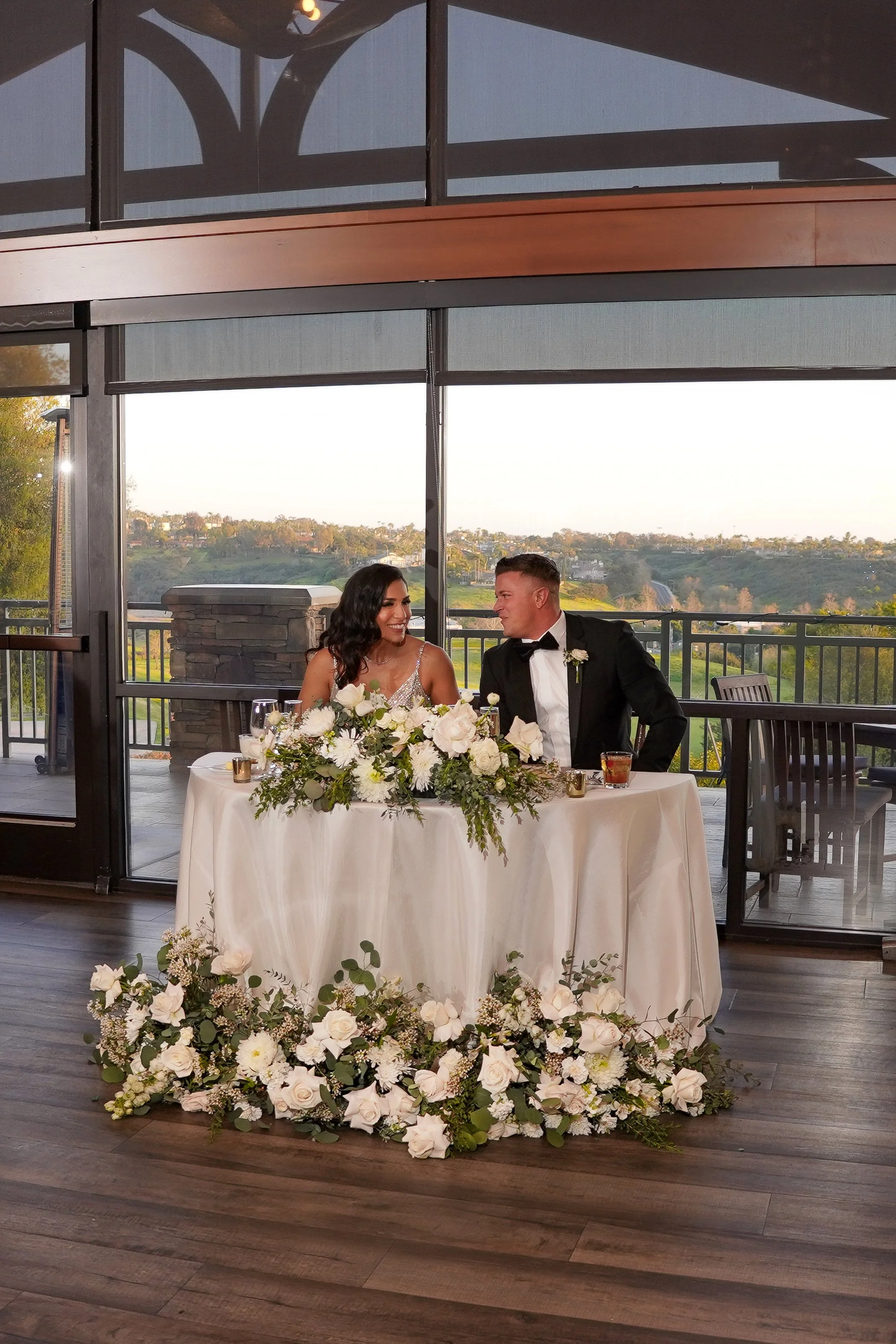 Wedding sweetheart table featuring a white floral centerpiece and floor-length flower arrangements, set against a sunset view through large windows at the Crossing Carlsbad reception.