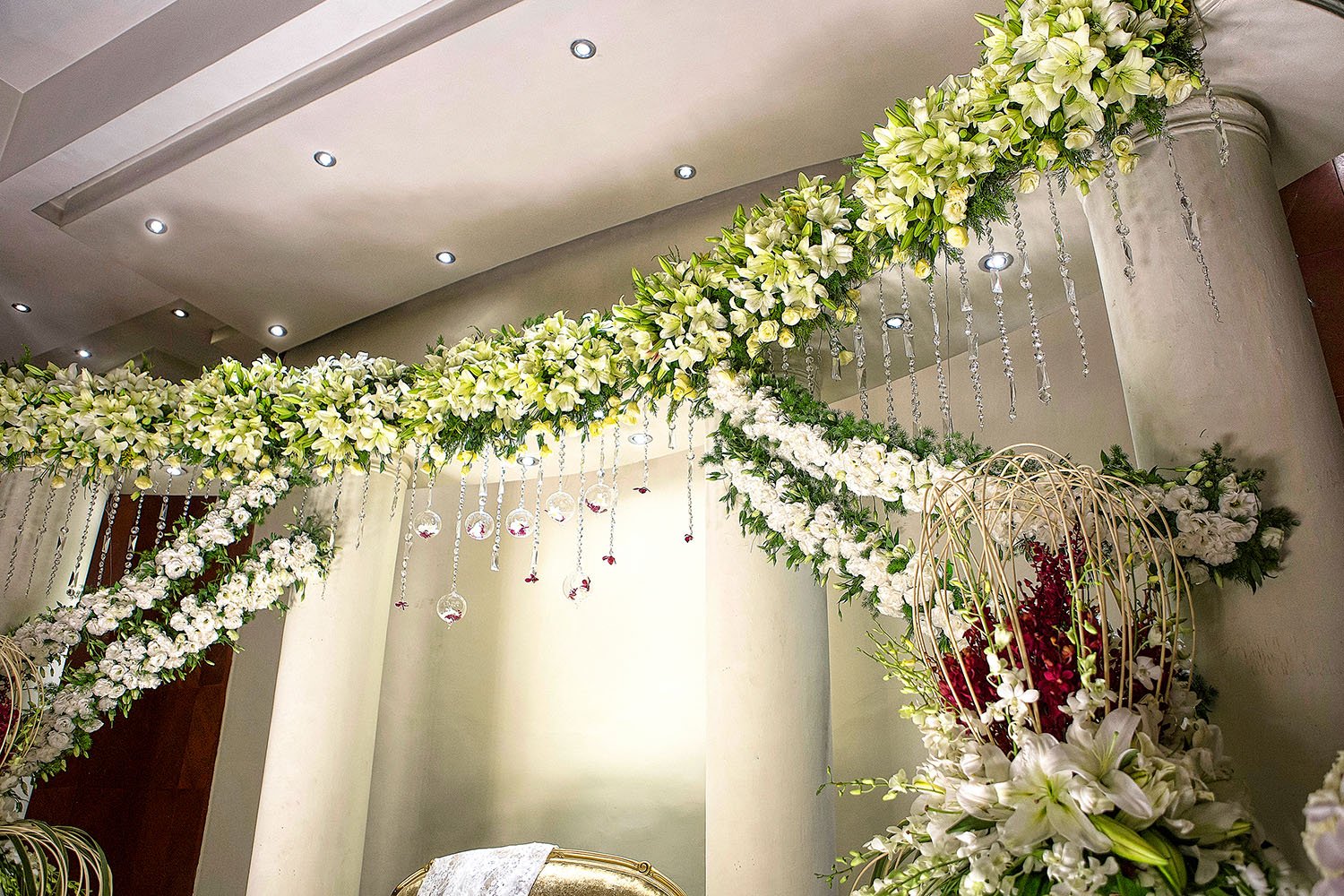 An elaborate indoor event floral display featuring large swags of white and green flowers (lilies, hydrangeas, orchids) draped across columns, with hanging crystal beads, clear glass baubles containing red accents, and a gold chair in the background.
