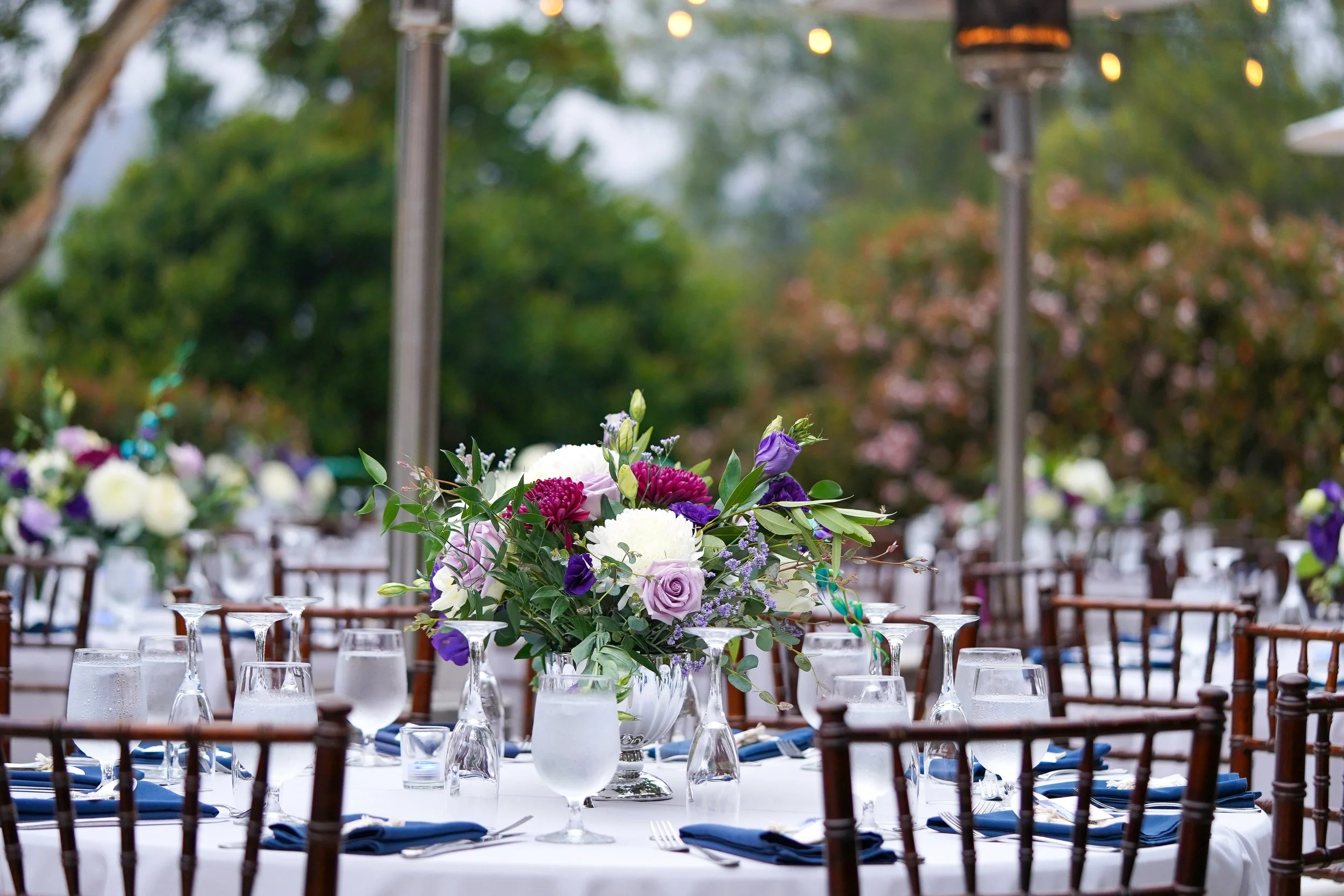 A round dining table set for an outdoor event with a white tablecloth, dark wood chairs, blue napkins, and a vibrant purple and white floral centerpiece under string lights.
