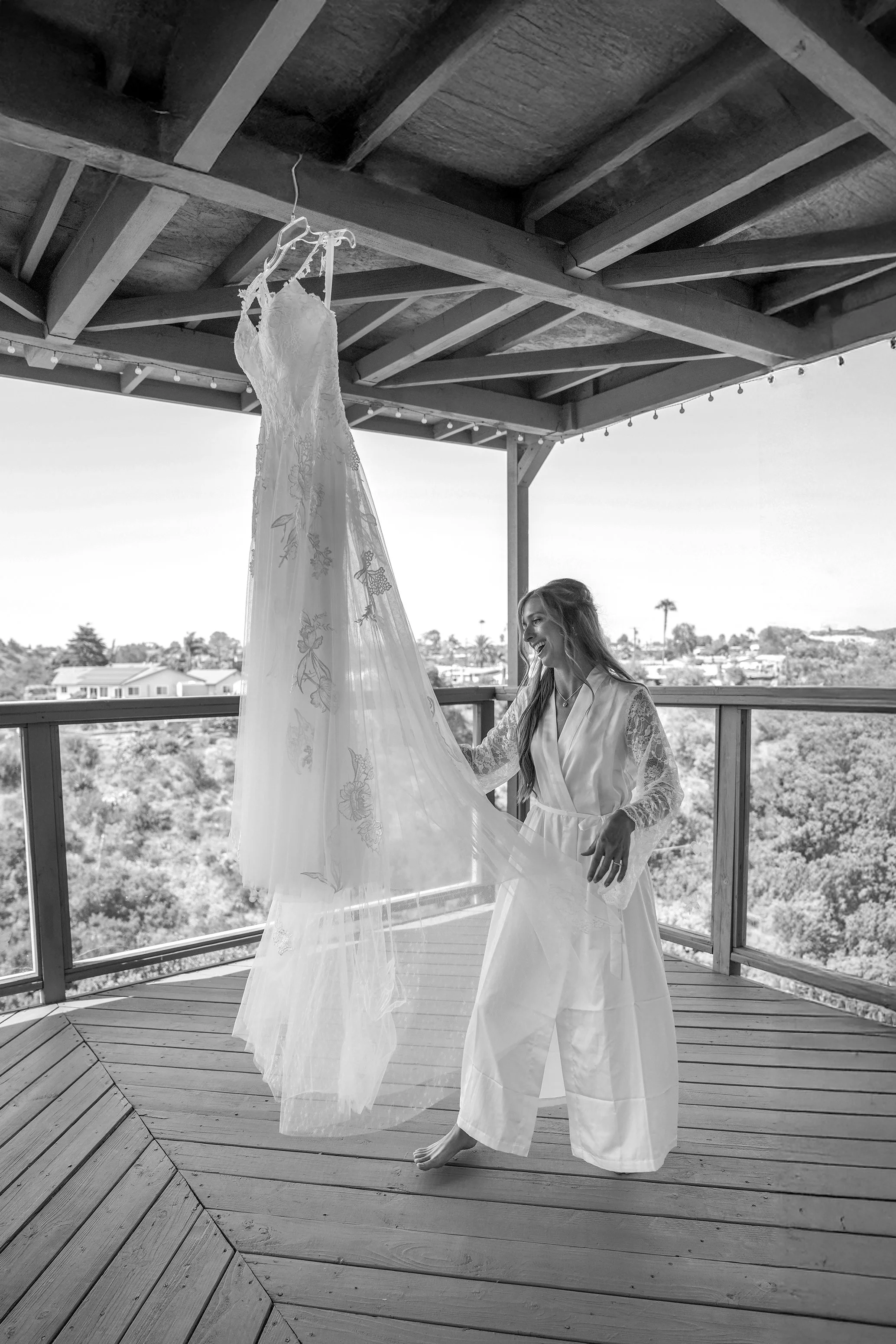 Bride joyfully touching her lace wedding gown hanging from a wooden porch during her wedding day preparations.